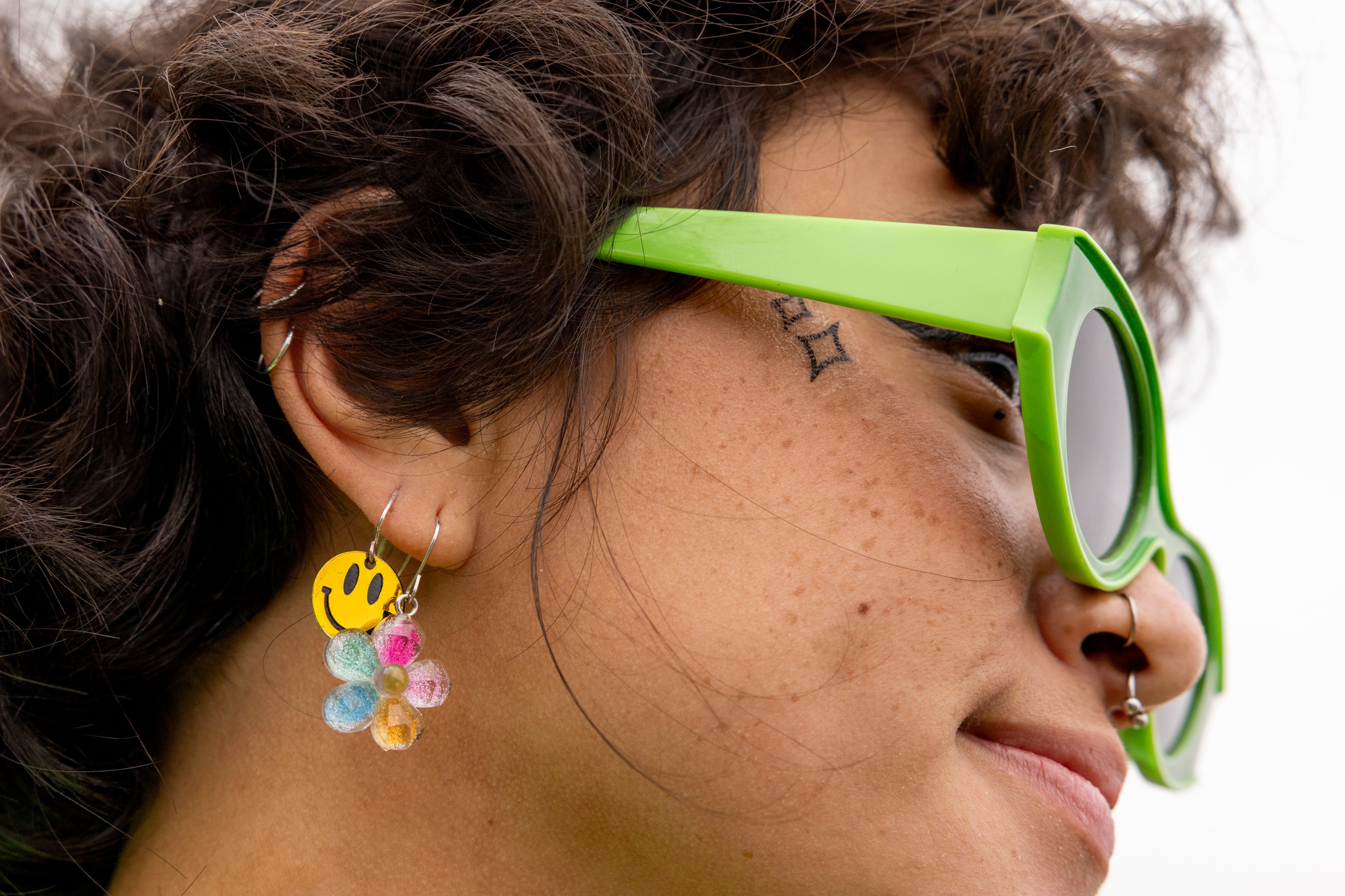 A person with wavy hair is wearing bright green sunglasses, smiley face earrings, and has a small diamond tattoo near their eye and a nose ring.