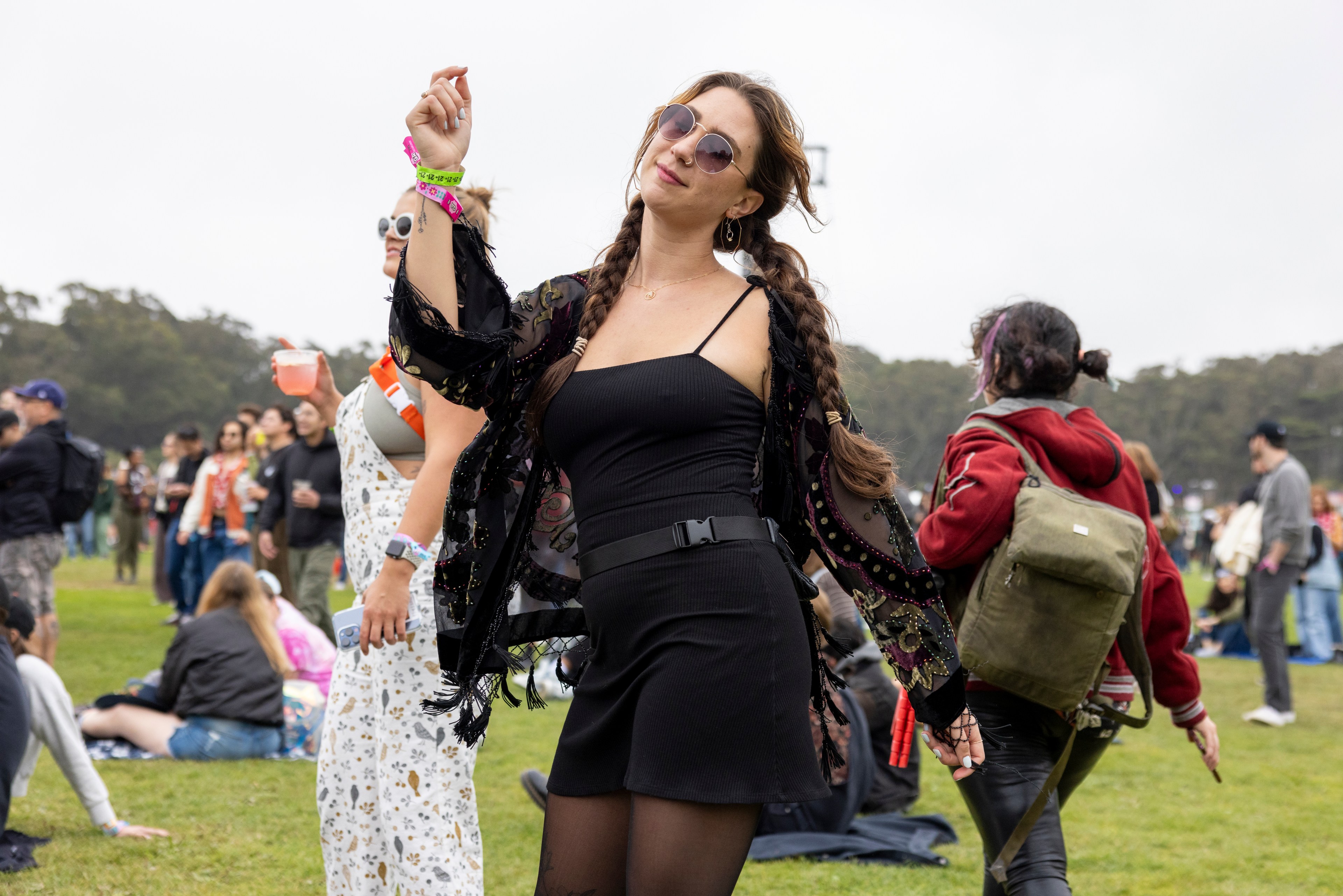 A woman in a black dress and sunglasses dances at an outdoor event, surrounded by others who are sitting and standing on a grassy field.