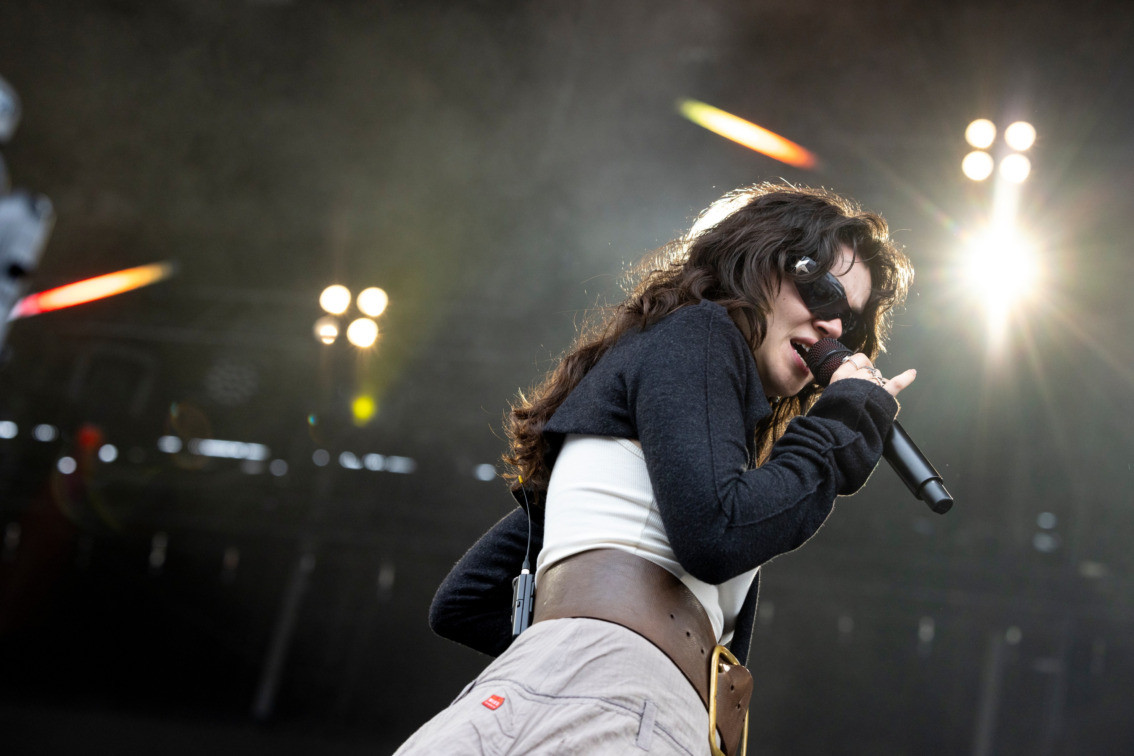 A performer with long hair and sunglasses sings into a microphone on stage, wearing a white top, dark sweater, and beige pants with a wide belt, under bright stage lights.