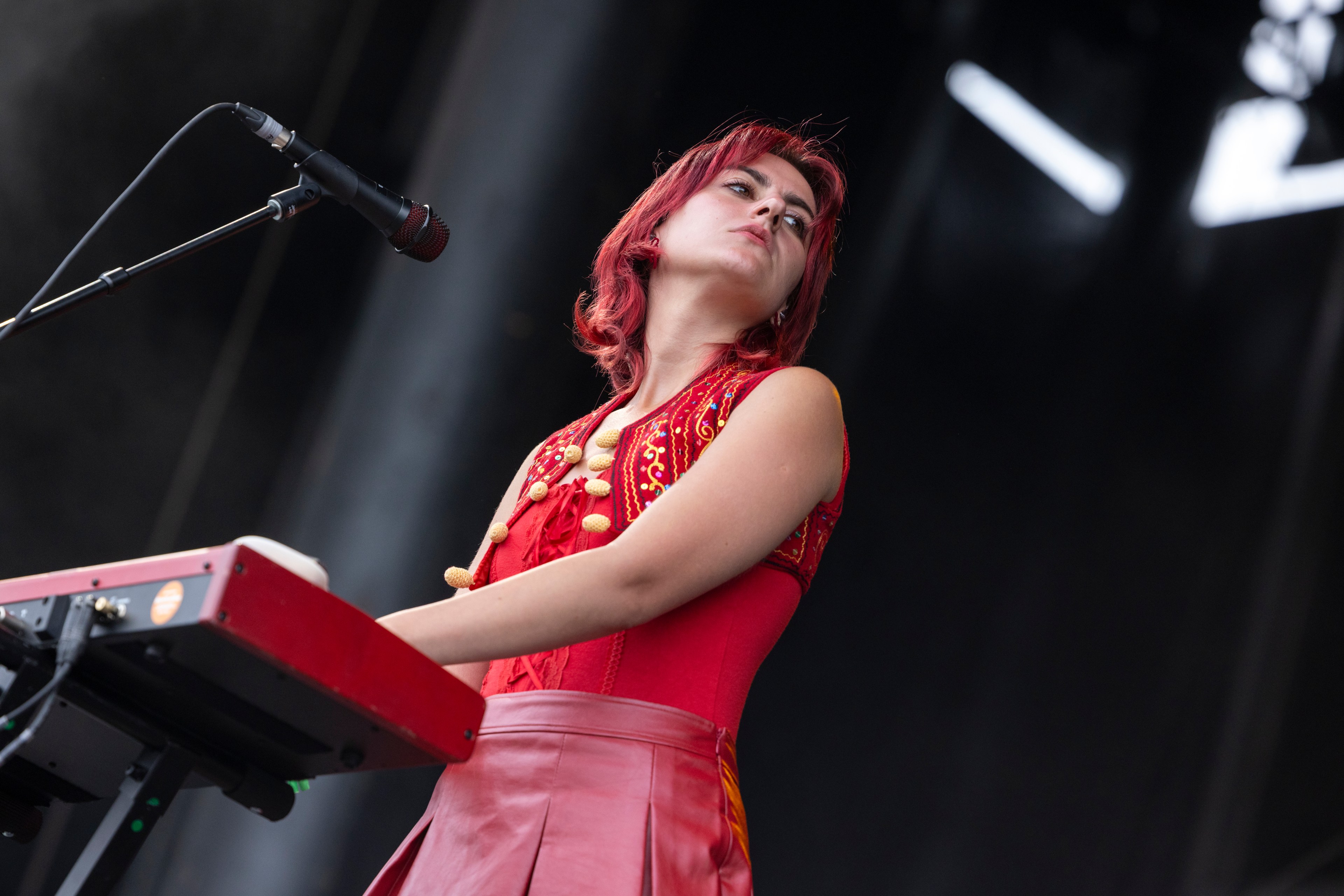 A person with red hair, wearing a red outfit, stands at a keyboard on stage, looking up with a serious expression. A microphone is positioned near their face.