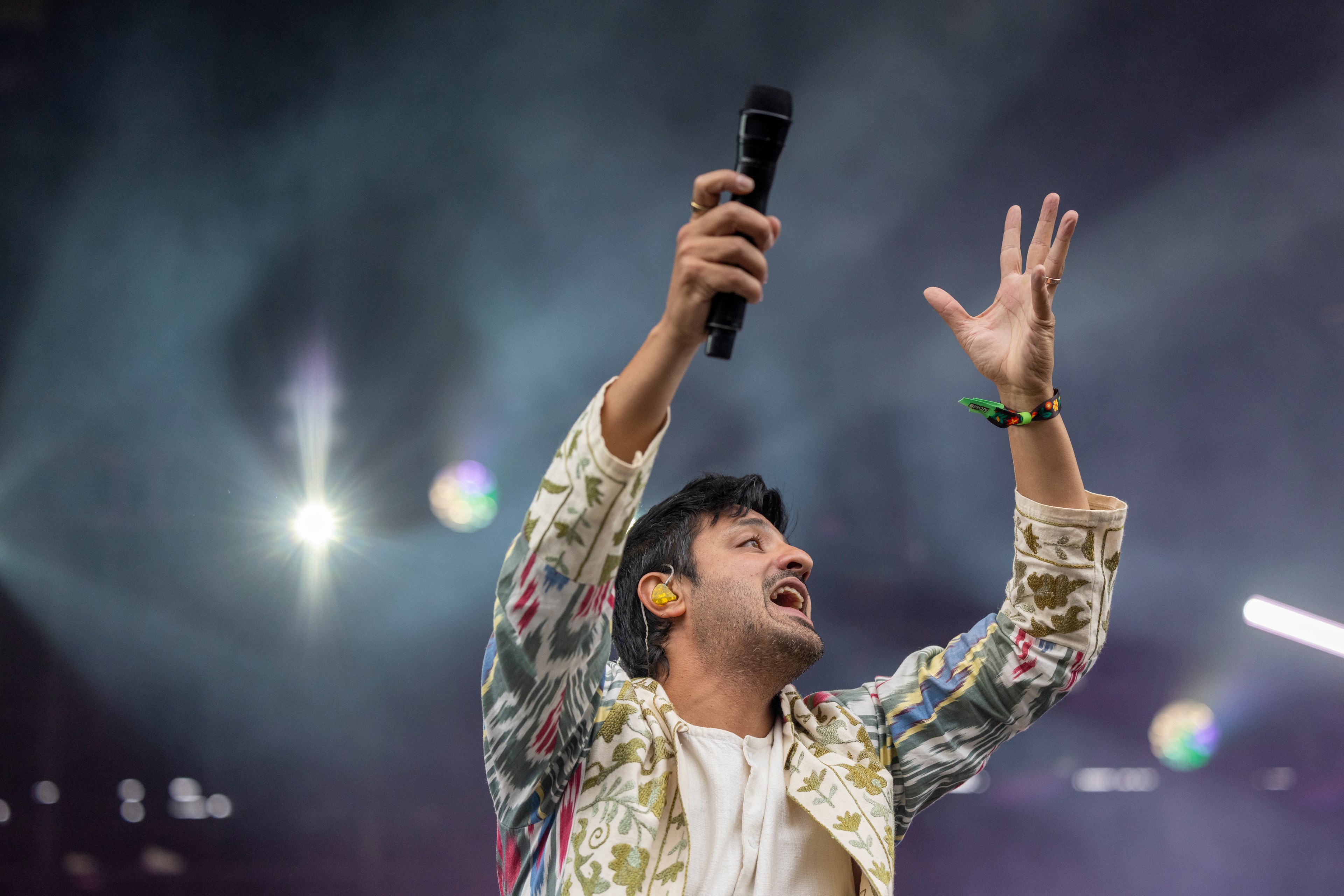 A man passionately sings into a microphone on stage, wearing a colorful patterned jacket, with dramatic stage lighting and smoke behind him.