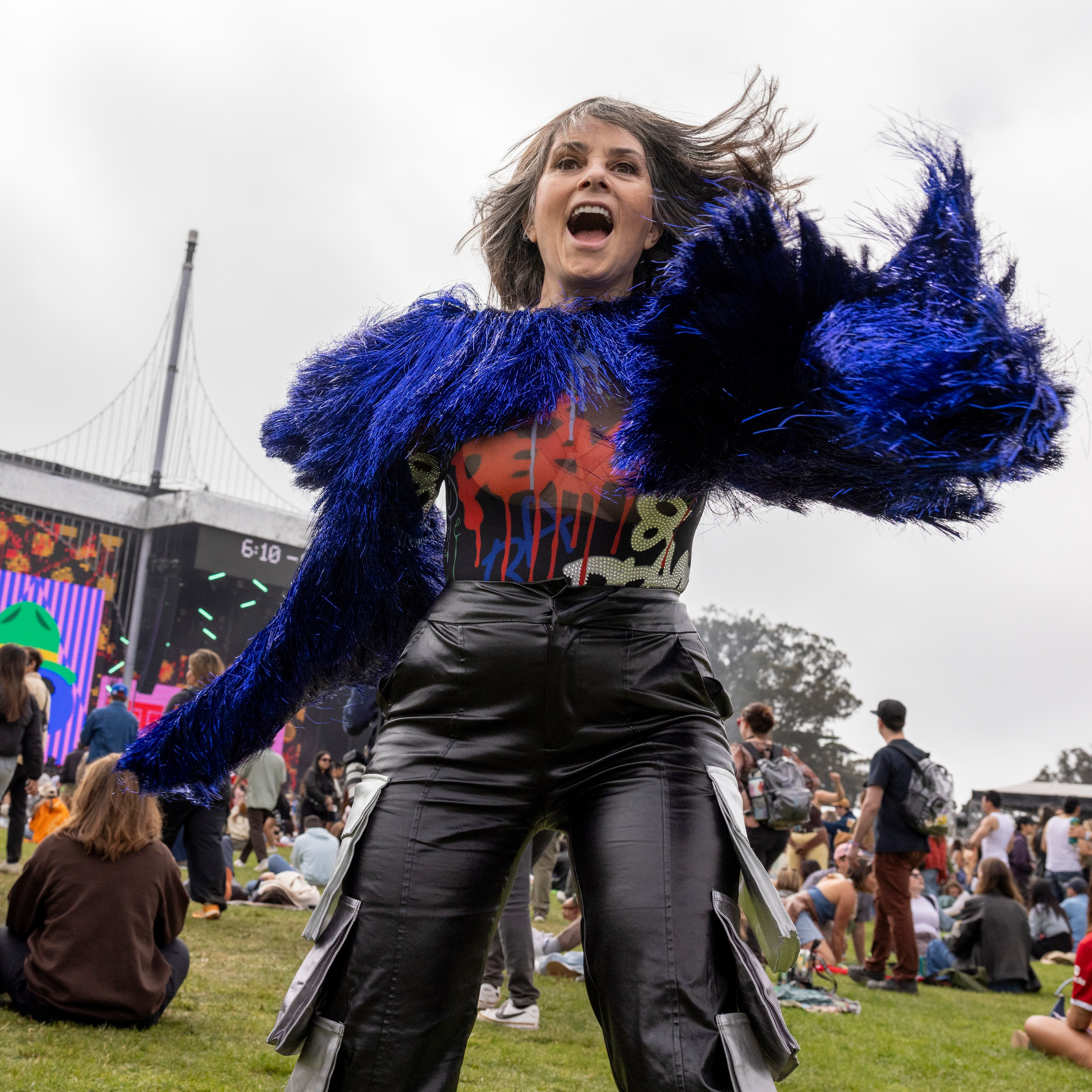 A woman in a vibrant blue, furry jacket dances energetically at an outdoor music festival. The background shows a large crowd and a lit stage with colorful screens.