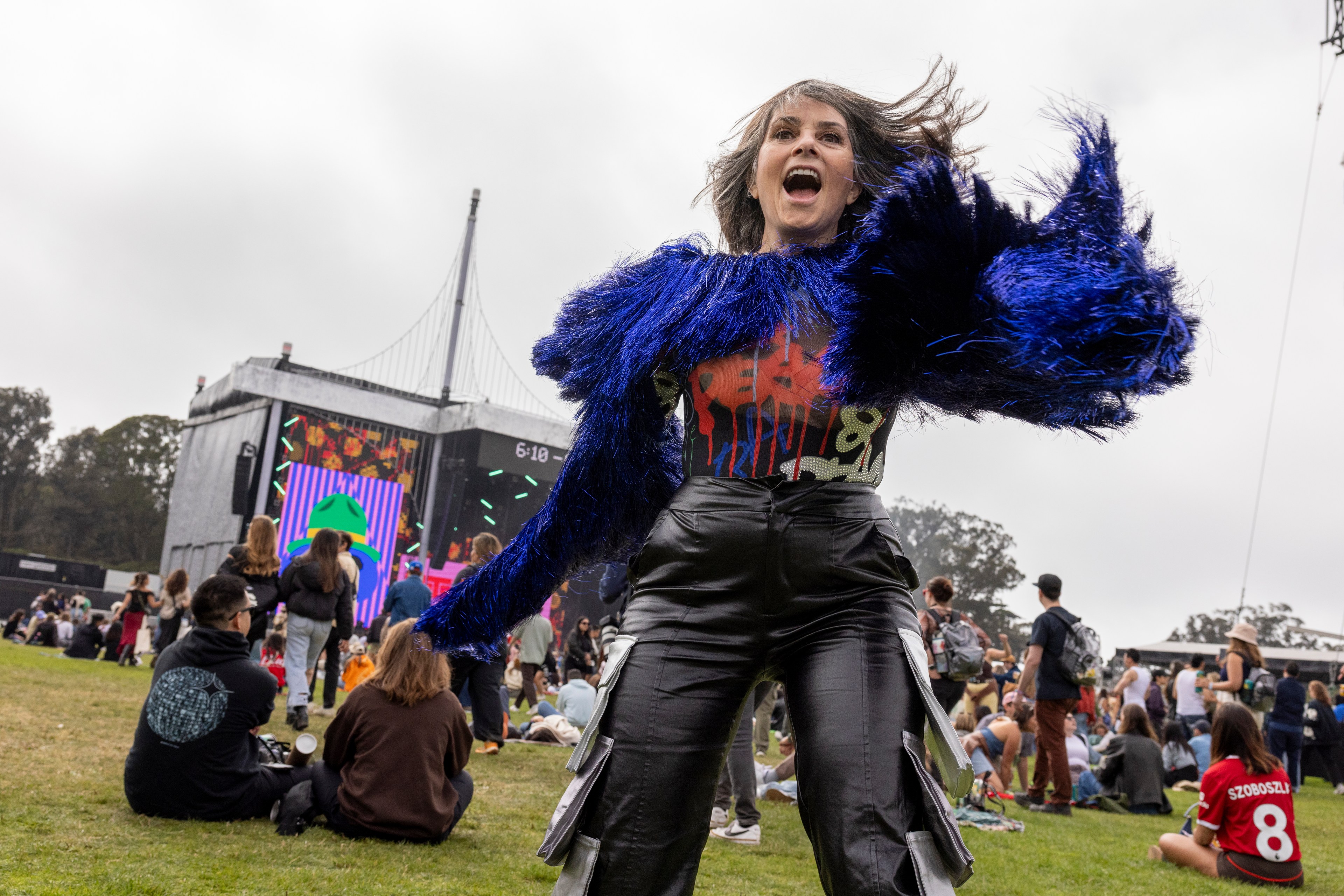 A woman in a vibrant blue, furry jacket dances energetically at an outdoor music festival. The background shows a large crowd and a lit stage with colorful screens.