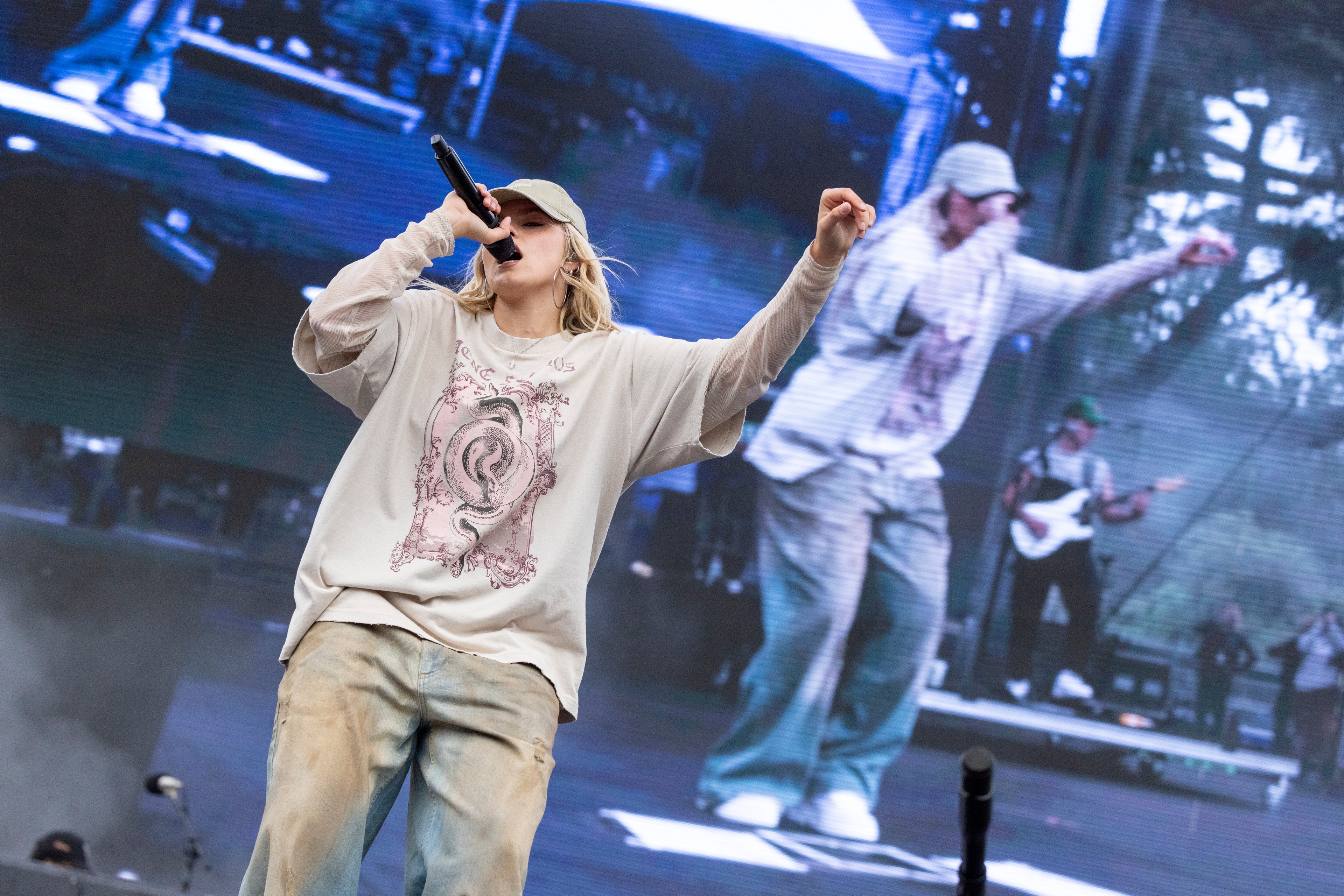 A person wearing a cap, long-sleeved shirt, and jeans performs on stage holding a microphone, with a larger screen in the background displaying their image.