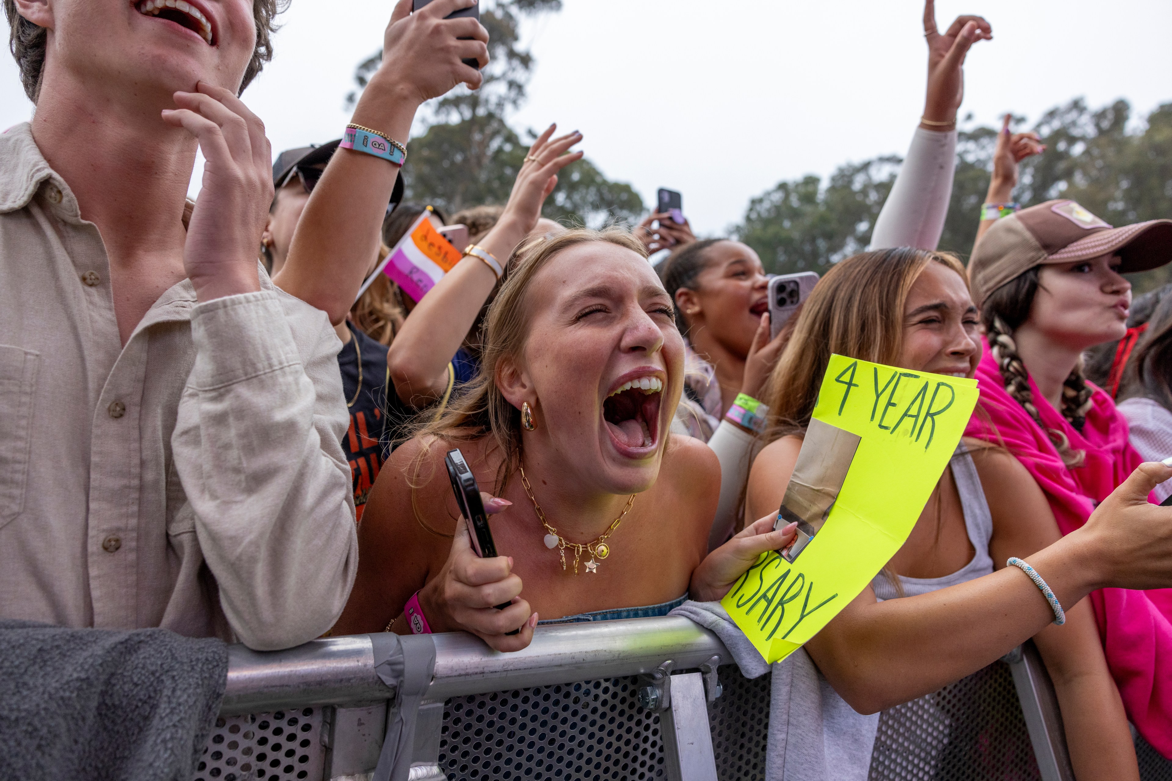 A group of excited people is cheering at a concert, with a woman in the center passionately yelling. One holds a bright yellow sign reading &quot;4 YEAR&quot; along a barricade.