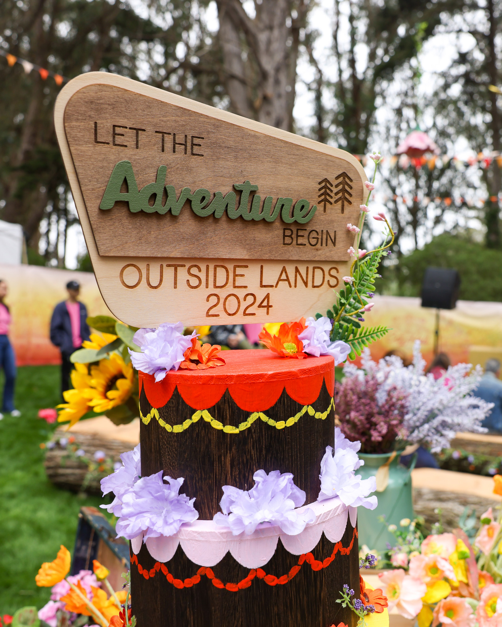 The image shows a wooden sign reading &quot;Let the Adventure Begin Outside Lands 2024,&quot; adorned with colorful flowers in an outdoor setting with people in the background.