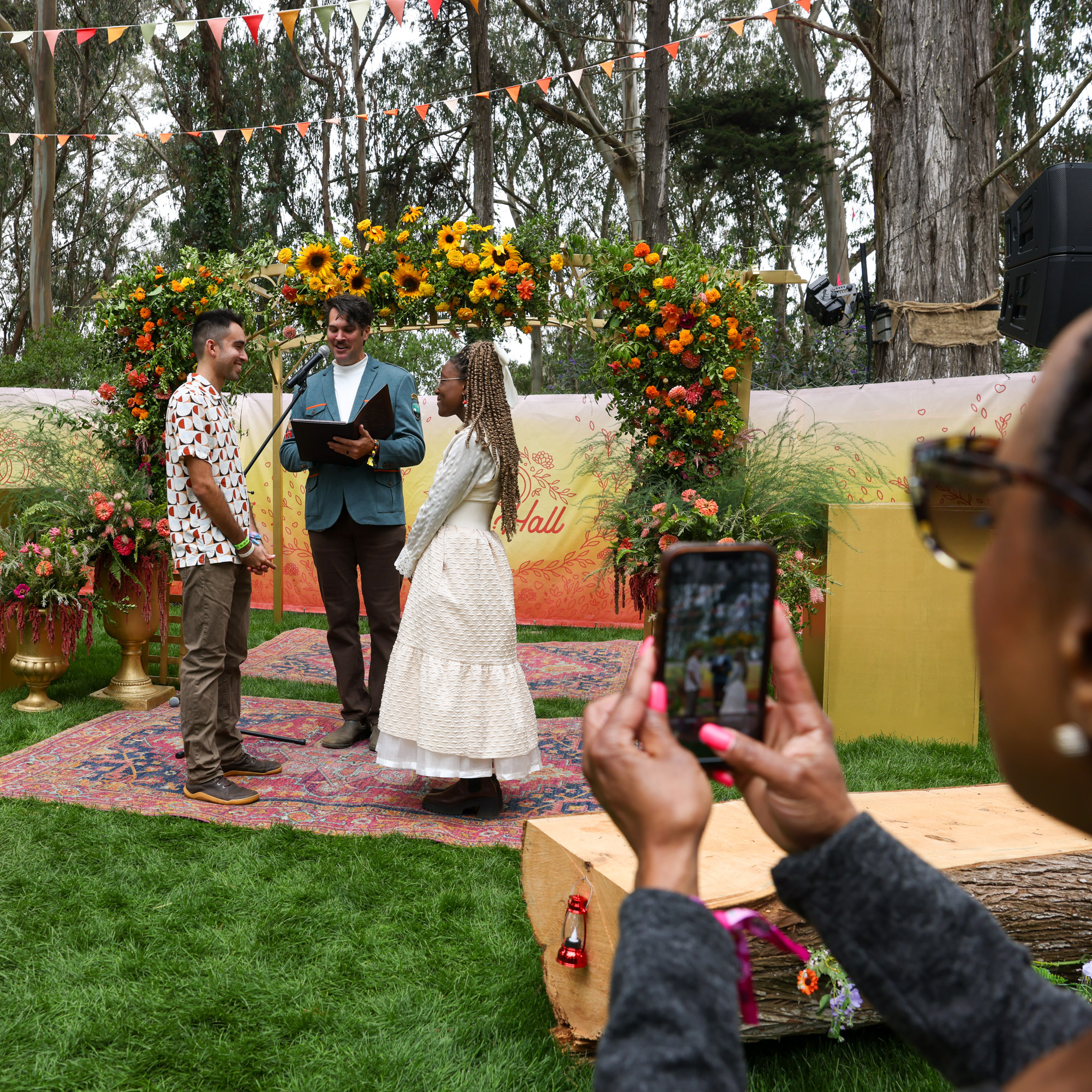 A couple stands facing each other and holding hands, while a person officiates their ceremony under a flower arch. Another person captures the moment on a smartphone.