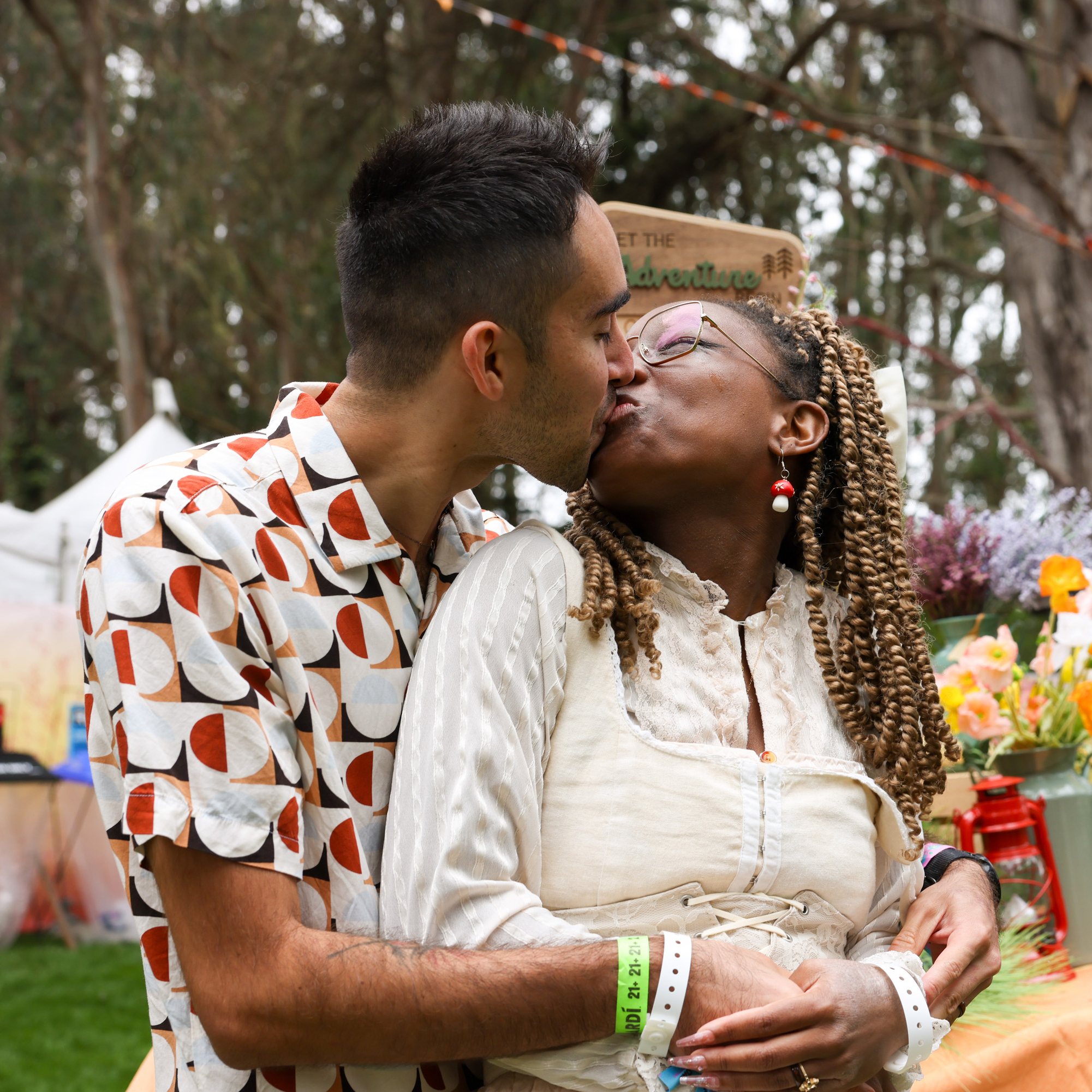 A couple stands outdoors, sharing a kiss. One wears a patterned shirt, the other a light dress and glasses. There are flowers and some fair-like decorations around them.