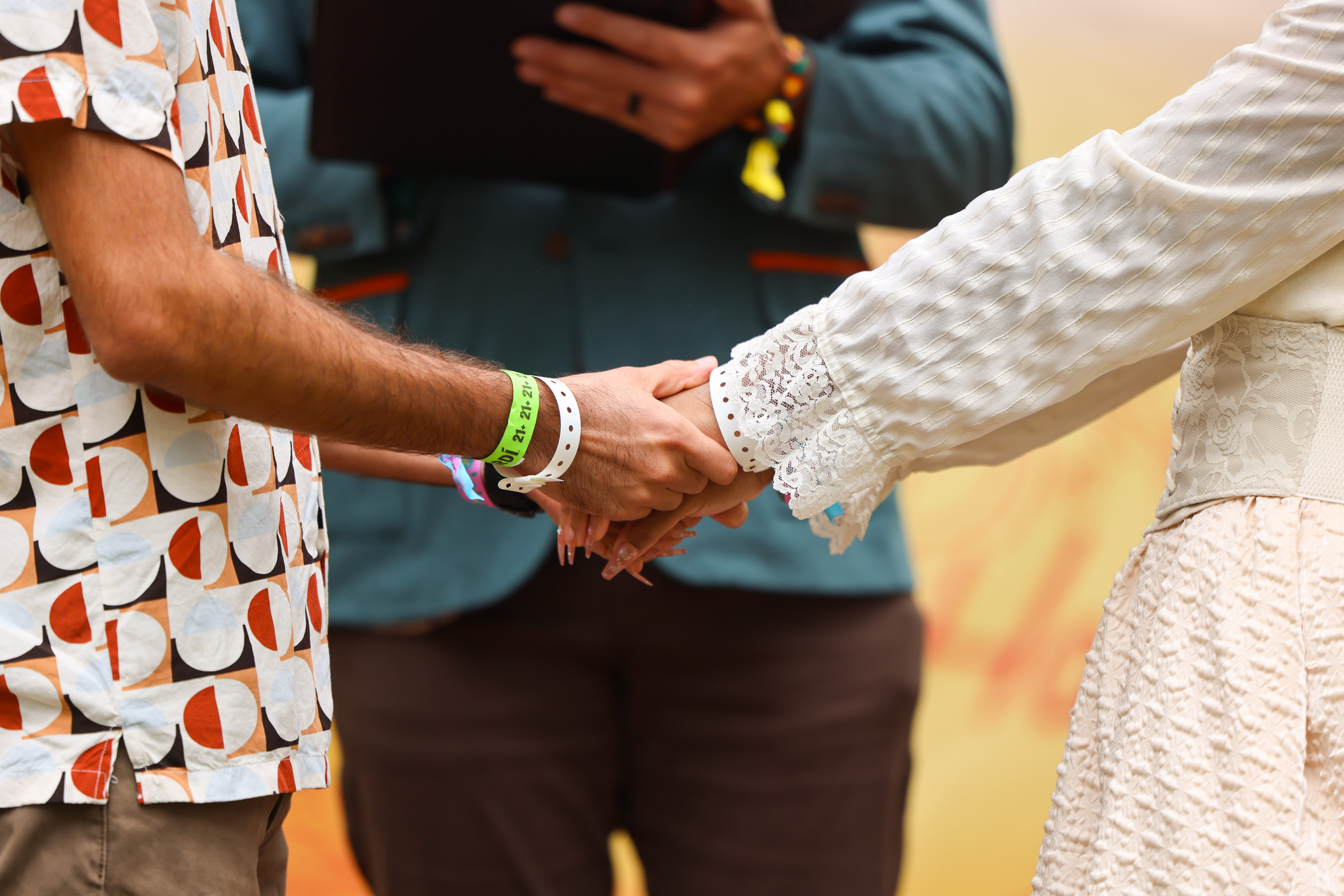 Two people are holding hands, one wearing a patterned shirt and the other a lace-trimmed sleeve dress, with an officiant holding a book in the background.