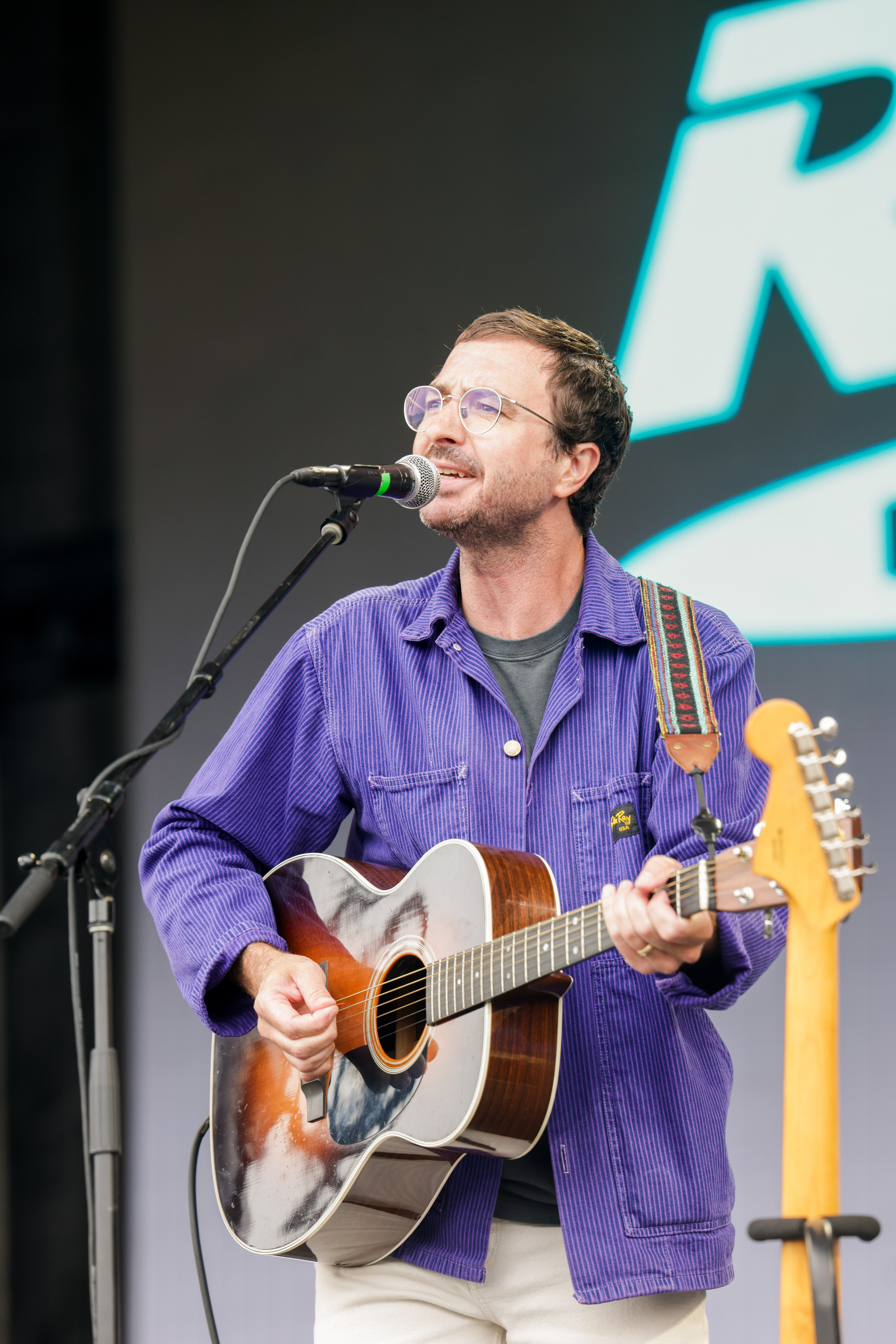 A person wearing glasses and a purple jacket plays an acoustic guitar and sings into a microphone on stage. There's a large logo in the background.