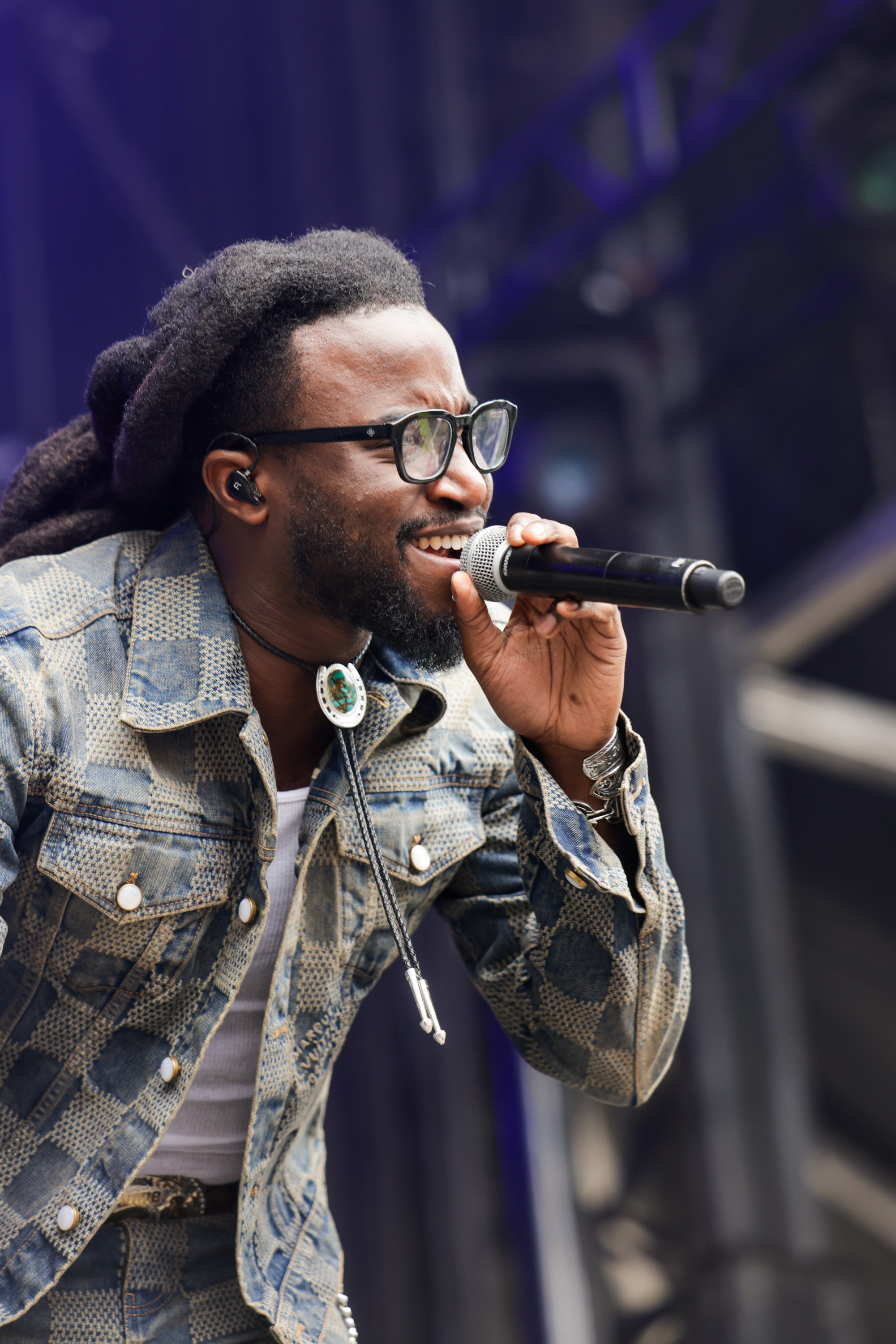 A man with long hair and glasses sings passionately into a microphone. He is dressed in a patterned denim jacket and wears a bolo tie.