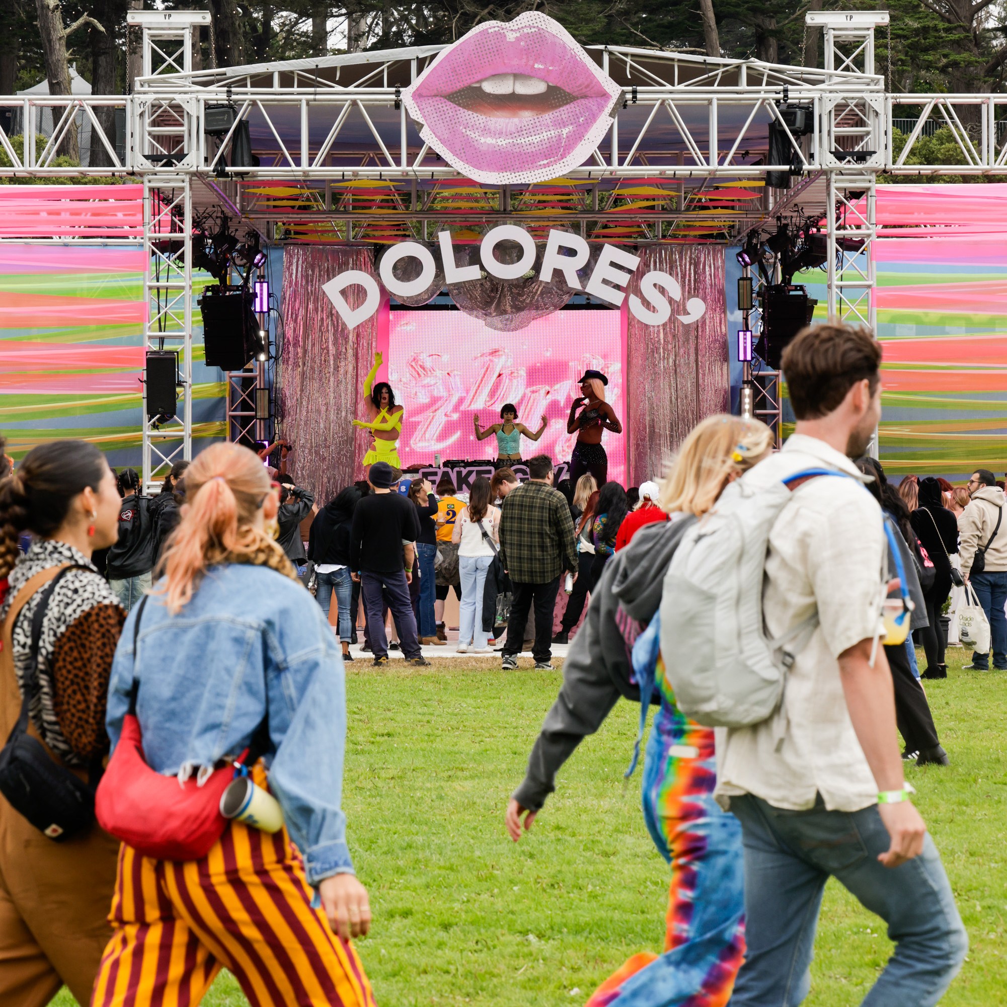 People gather at an outdoor festival stage with a large pink lips decoration above and the word &quot;DOLORES&quot; displayed, while performers entertain the crowd.