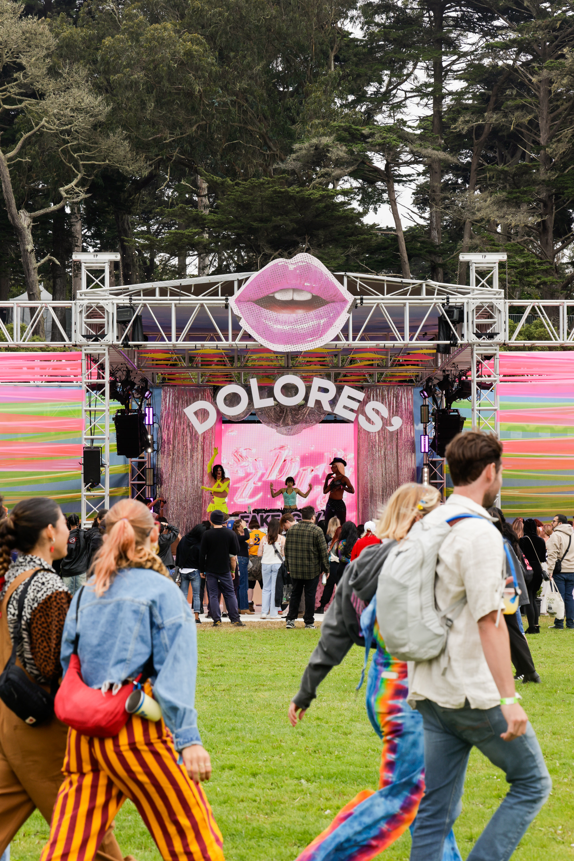 People gather at an outdoor festival stage with a large pink lips decoration above and the word &quot;DOLORES&quot; displayed, while performers entertain the crowd.