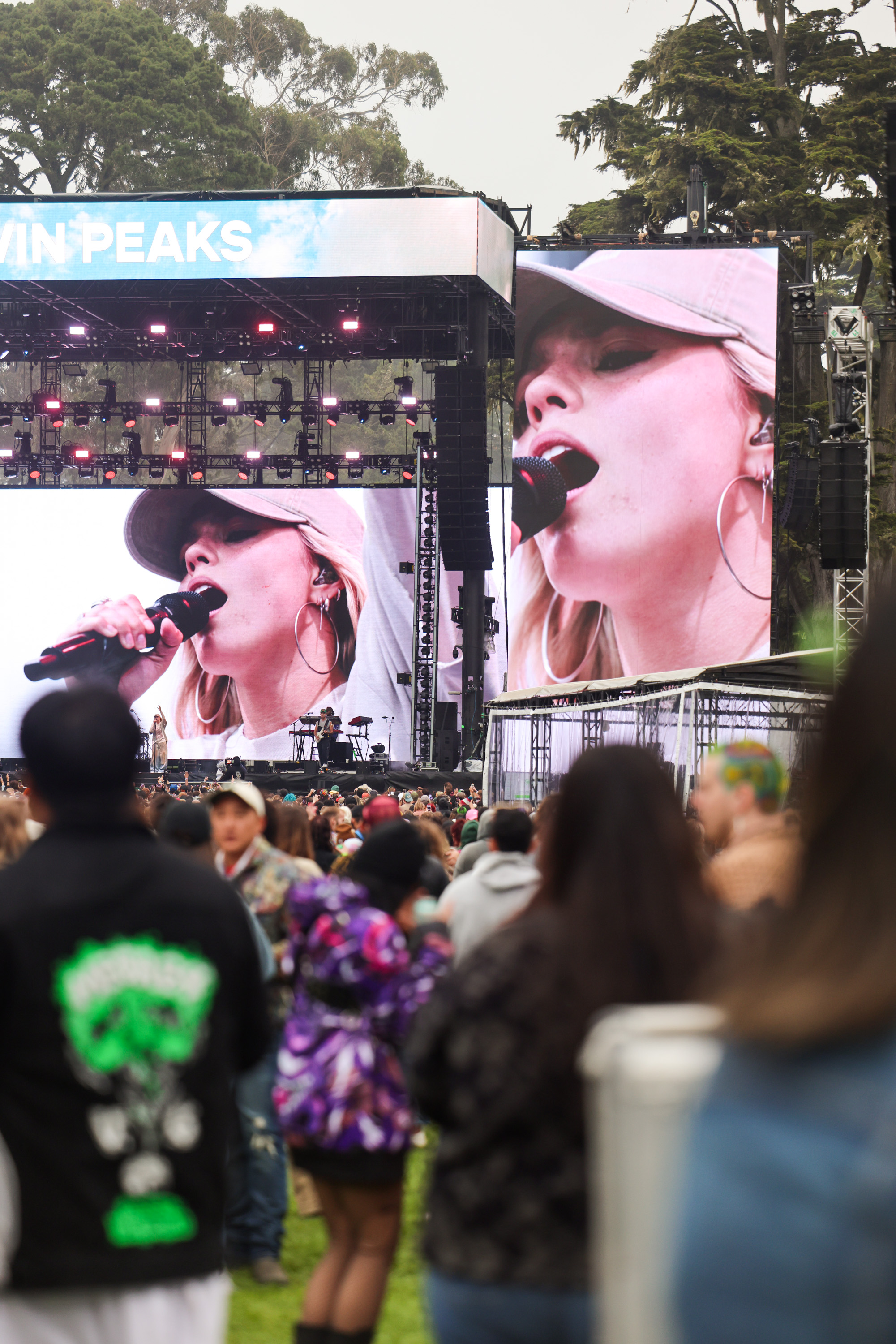 A large outdoor concert stage features a close-up of a female singer with a cap on a big screen, while a crowd of people watches. Trees are visible in the background.