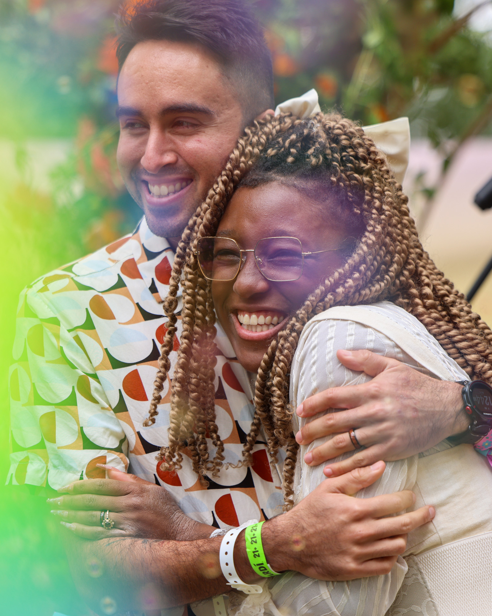 Two people are joyfully hugging. One smiles brightly, wearing glasses and colorful, braided hair. The other wears a patterned shirt, grinning while embracing tightly.