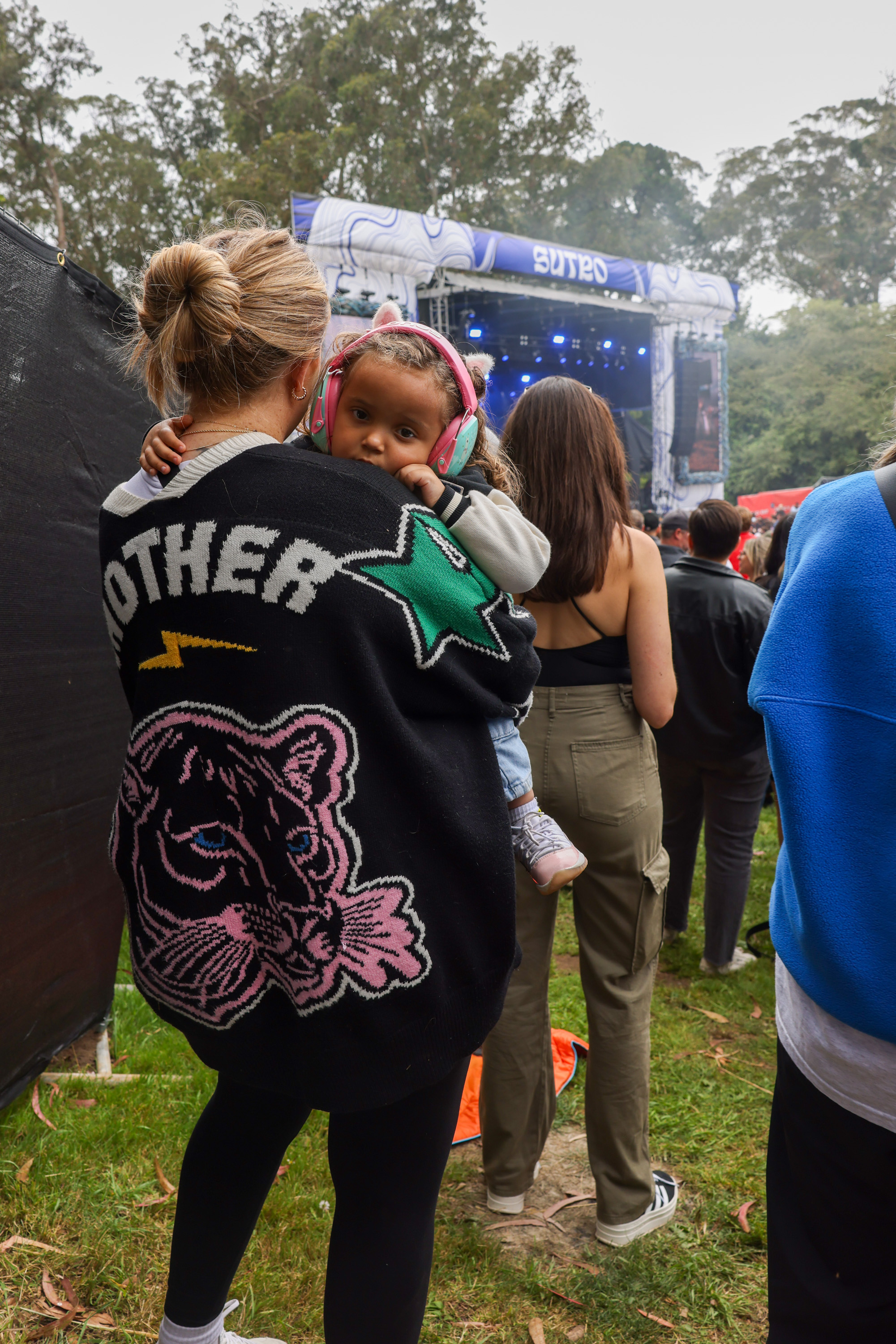 A woman holding a toddler wearing pink headphones at an outdoor concert, with a stage in the background surrounded by trees and other attendees.