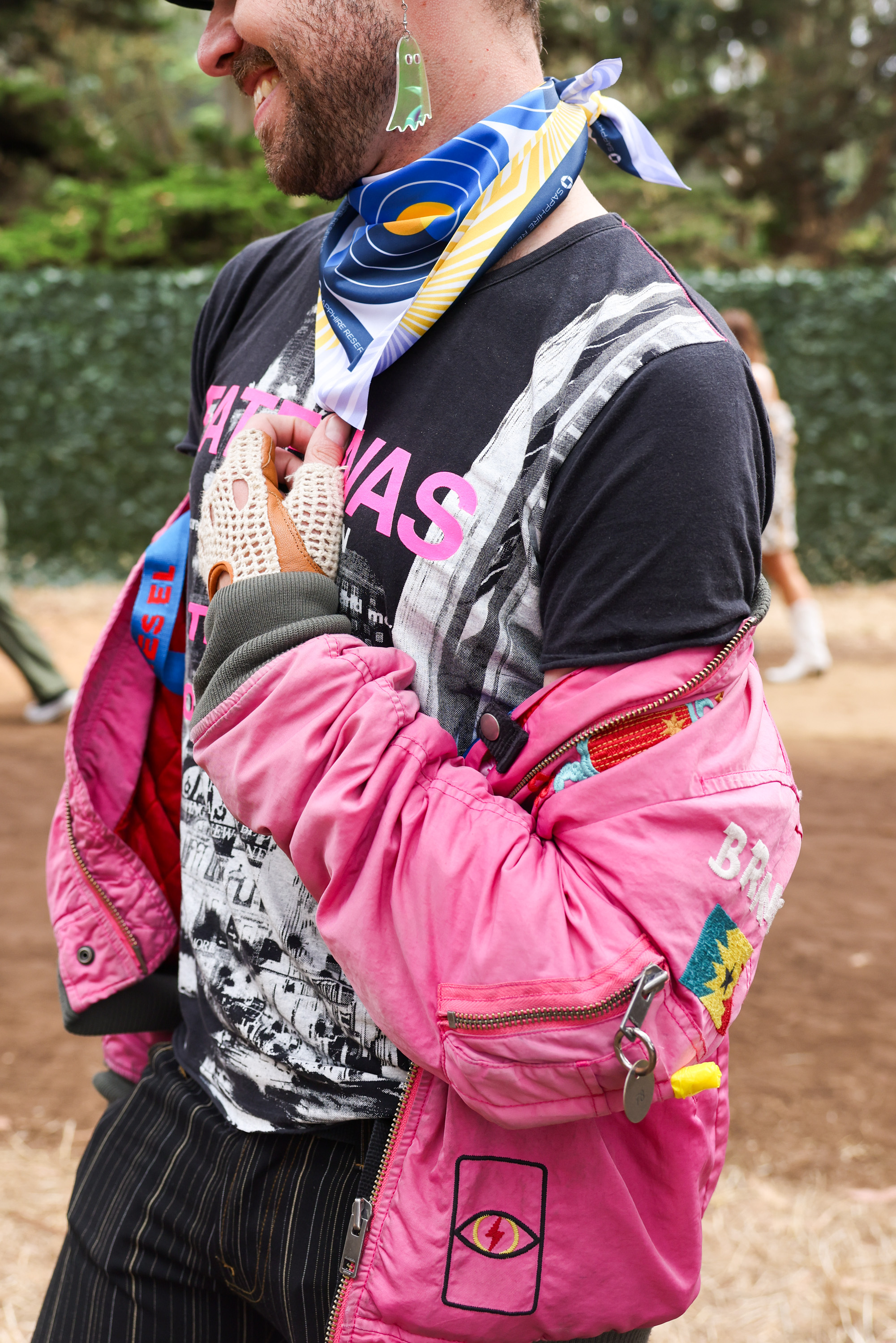 A person wears a pink jacket, black graphic tee, striped pants, a colorful scarf, and an earring shaped like a small ghost. They are smiling, partially obscured from view.