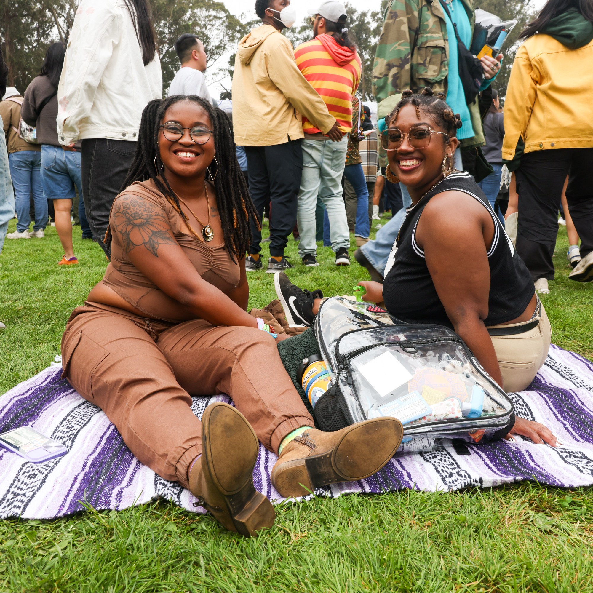 Two women sit on a blanket at an outdoor event, smiling at the camera, surrounded by a crowd of people. Both seem relaxed and happy, enjoying the atmosphere.