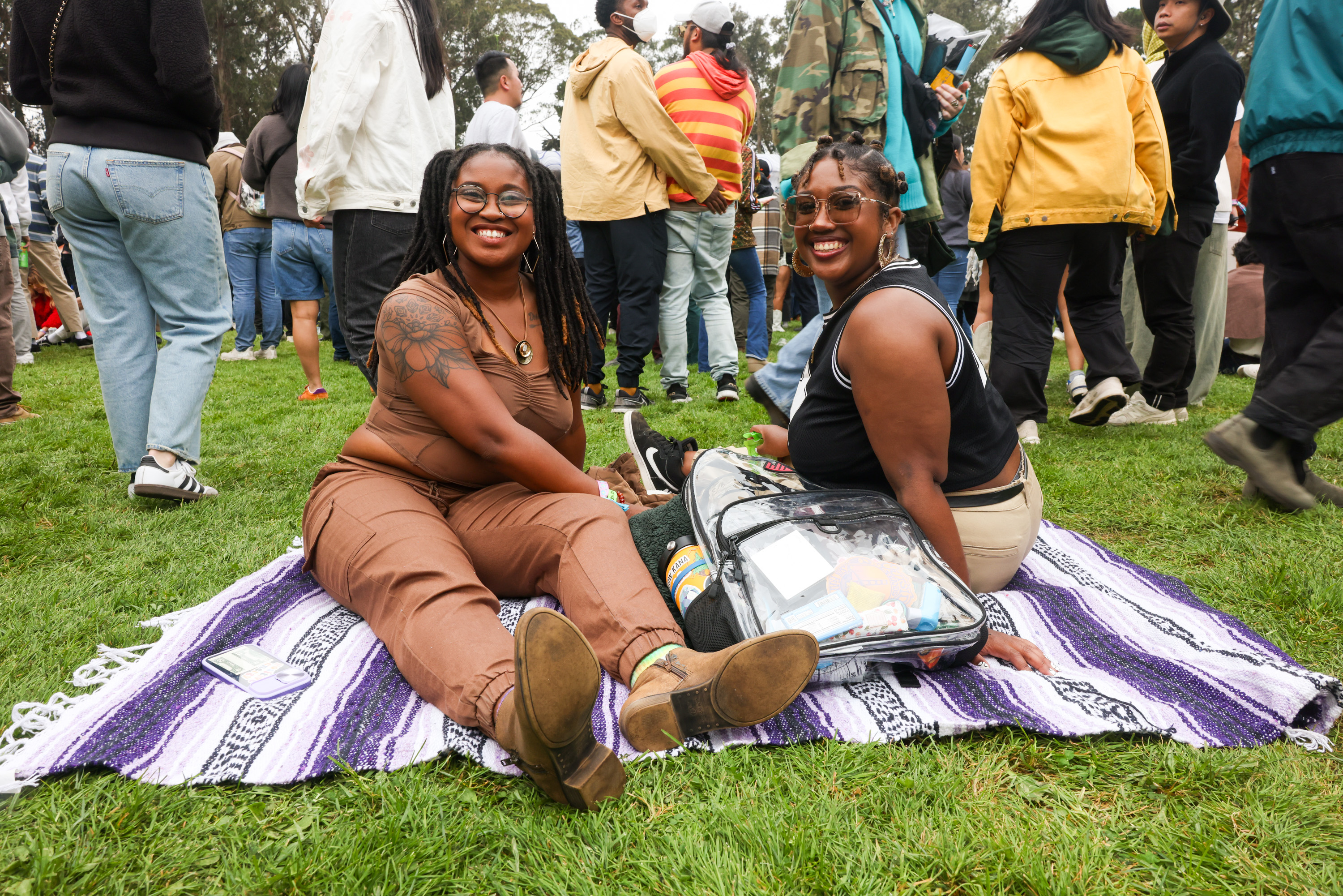 Two women sit on a blanket at an outdoor event, smiling at the camera, surrounded by a crowd of people. Both seem relaxed and happy, enjoying the atmosphere.