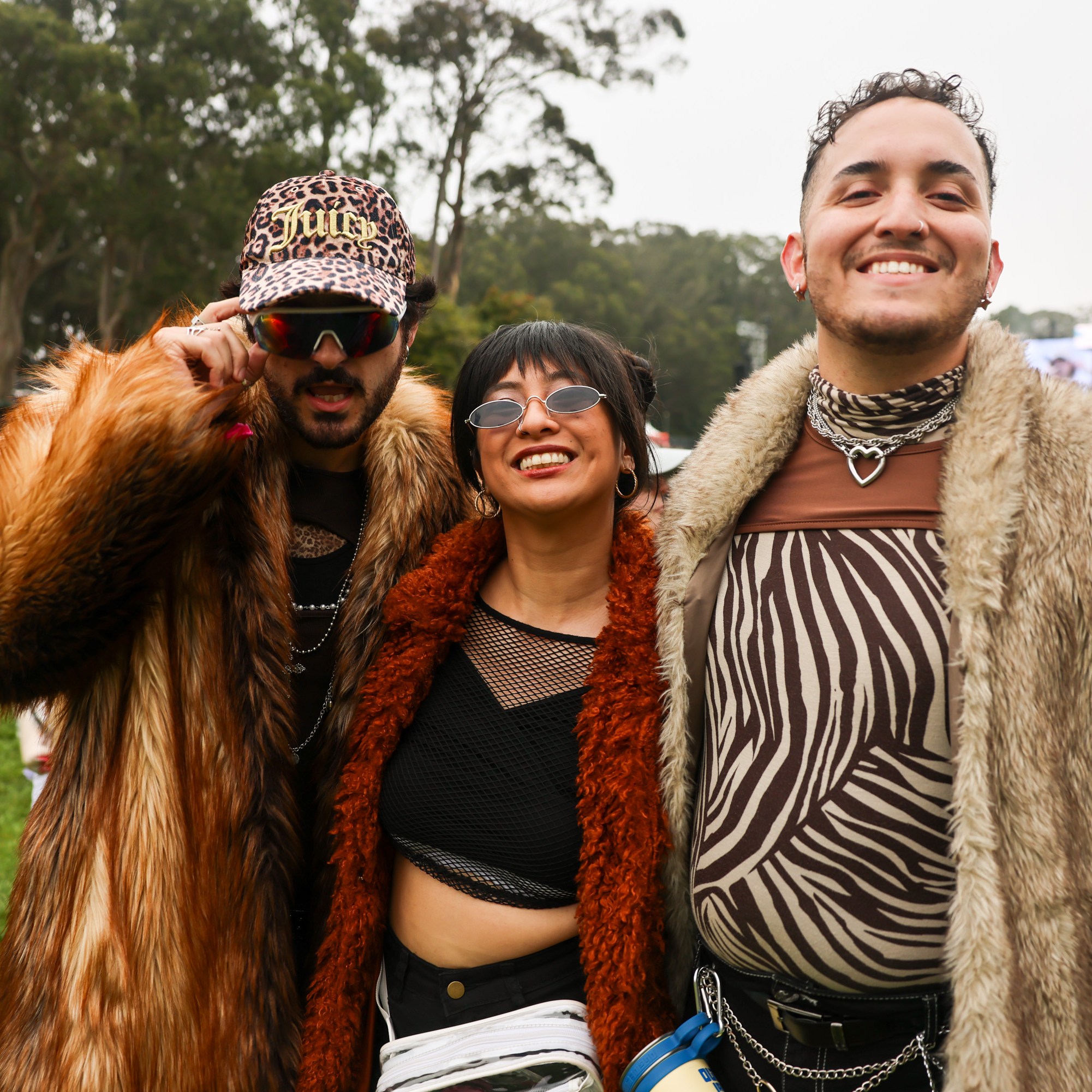 Three people are dressed in colorful, bold outfits, standing happily together outdoors. Trees and a large stage with screens in the background imply a festival setting.