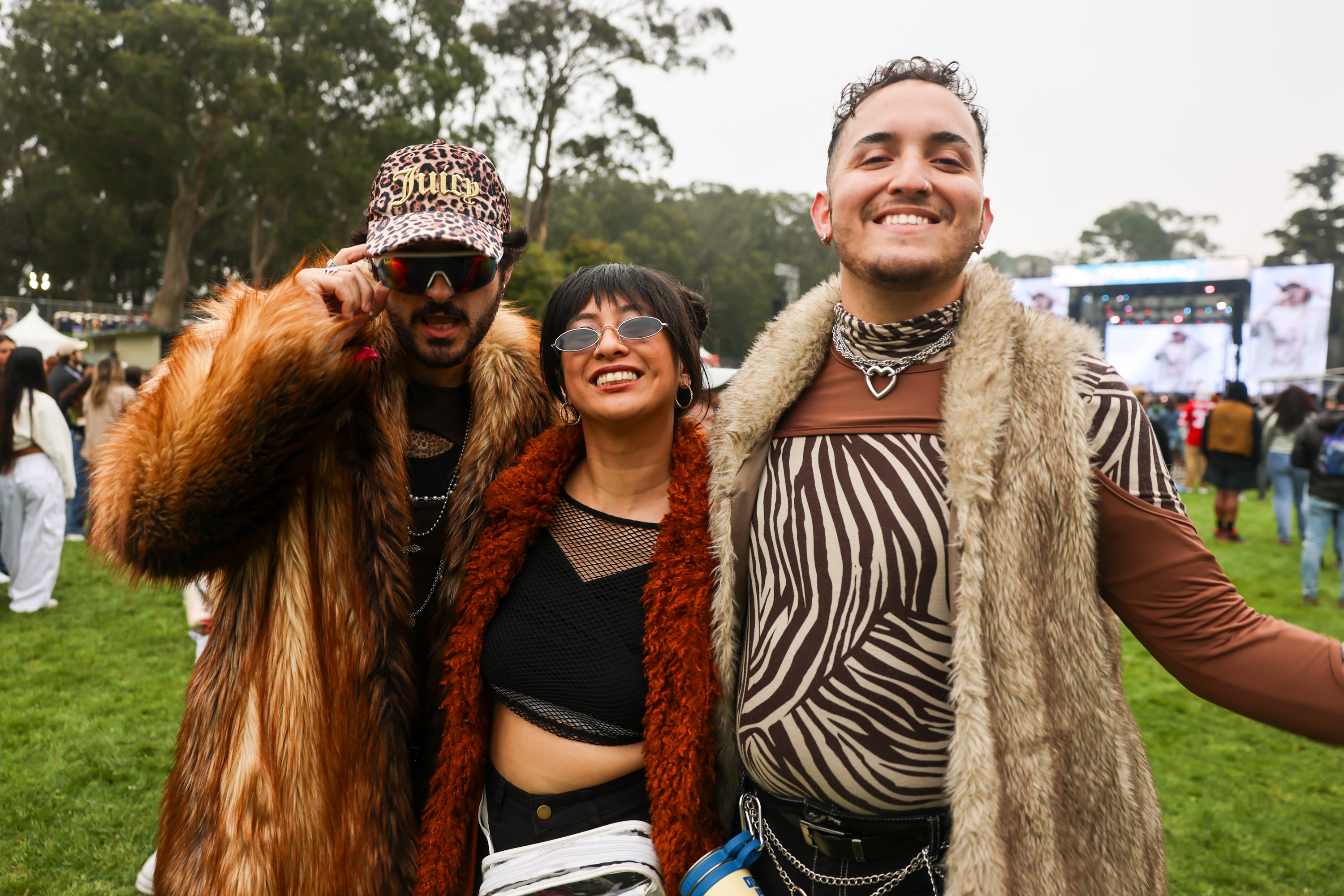 Three people are dressed in colorful, bold outfits, standing happily together outdoors. Trees and a large stage with screens in the background imply a festival setting.