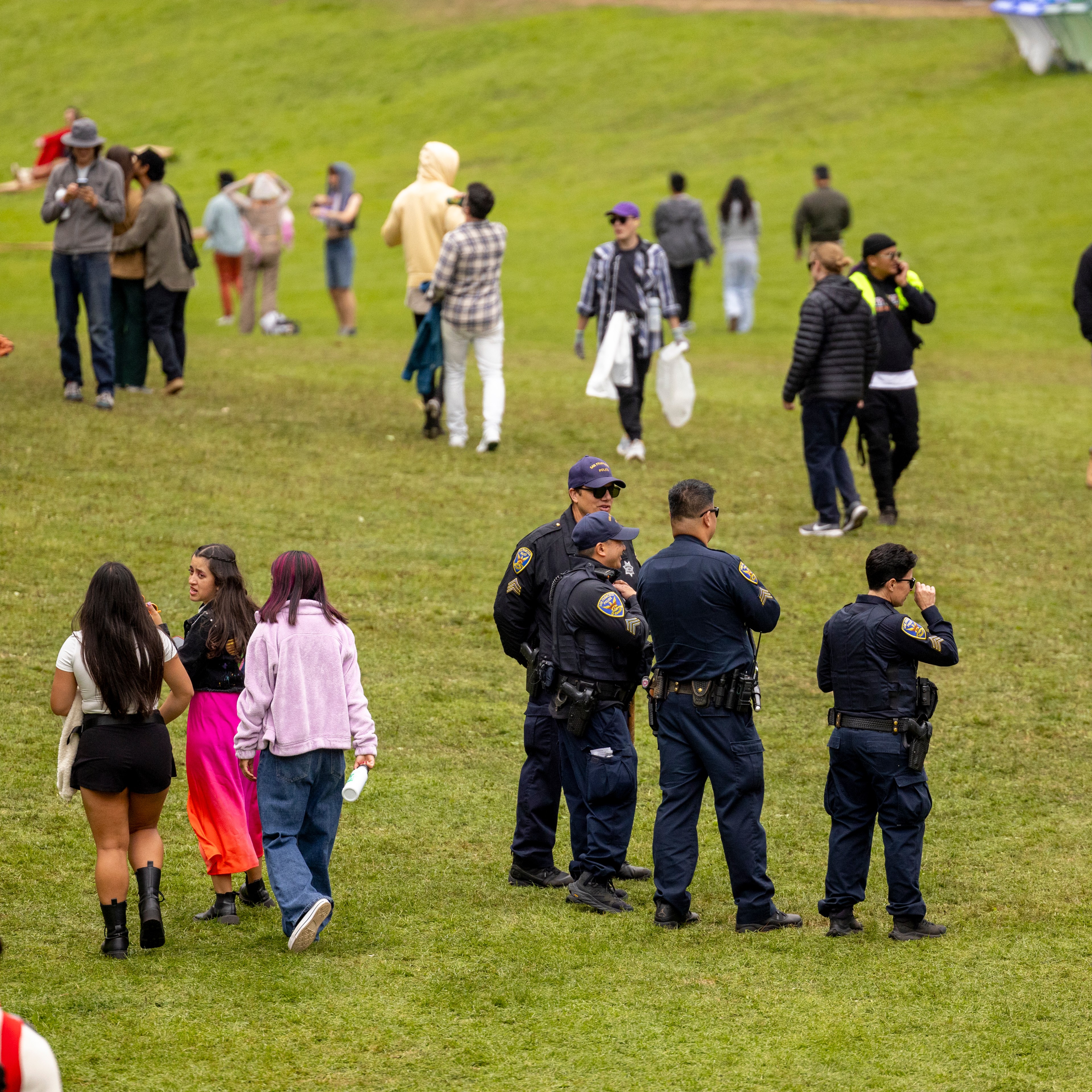 The image shows a grassy field with groups of people and several police officers. Some are walking, others are standing and chatting. The scene is casual and relaxed.