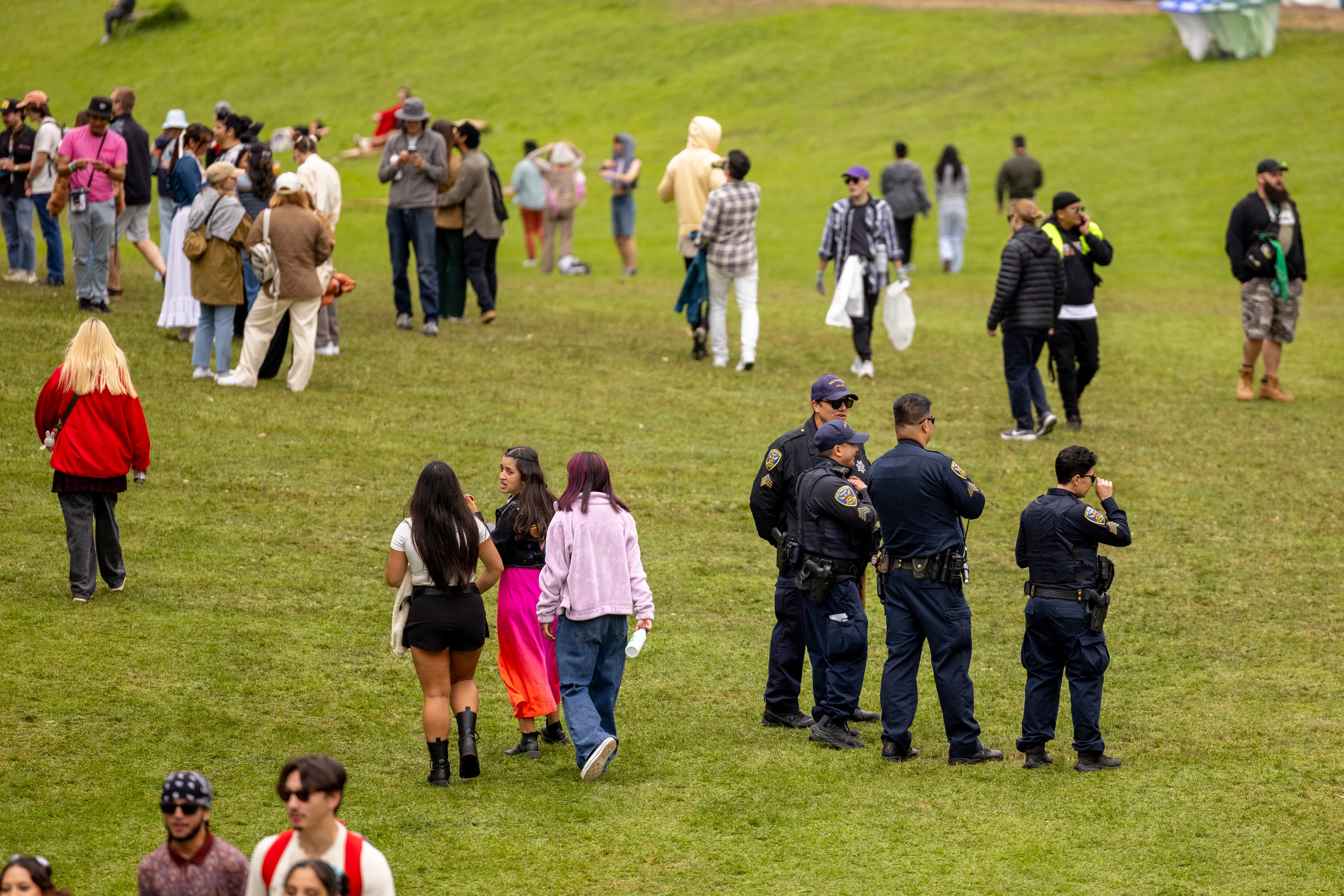 The image shows a grassy field with groups of people and several police officers. Some are walking, others are standing and chatting. The scene is casual and relaxed.