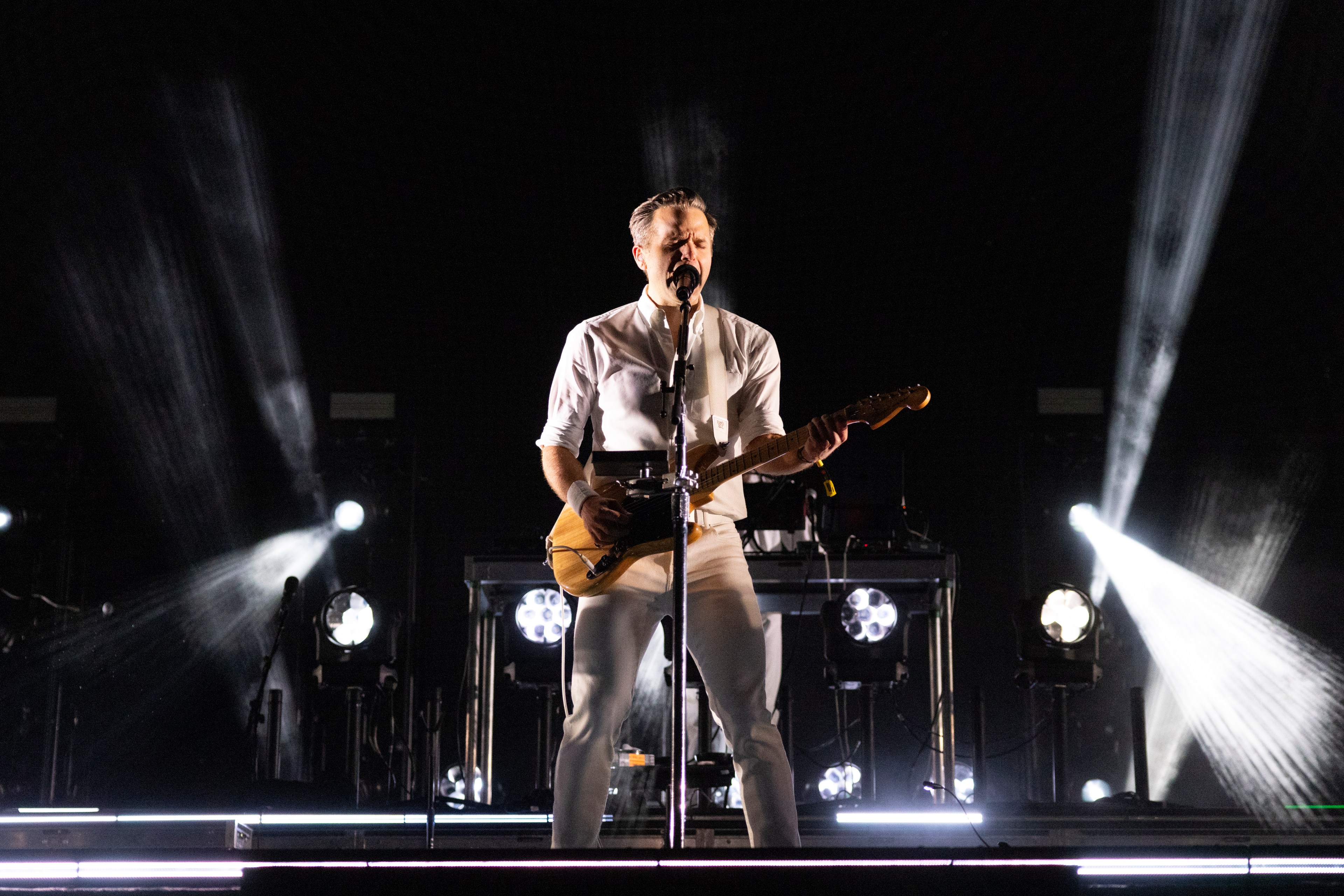 A musician in white clothing passionately sings into a microphone while playing an electric guitar onstage, with bright spotlights illuminating the scene.