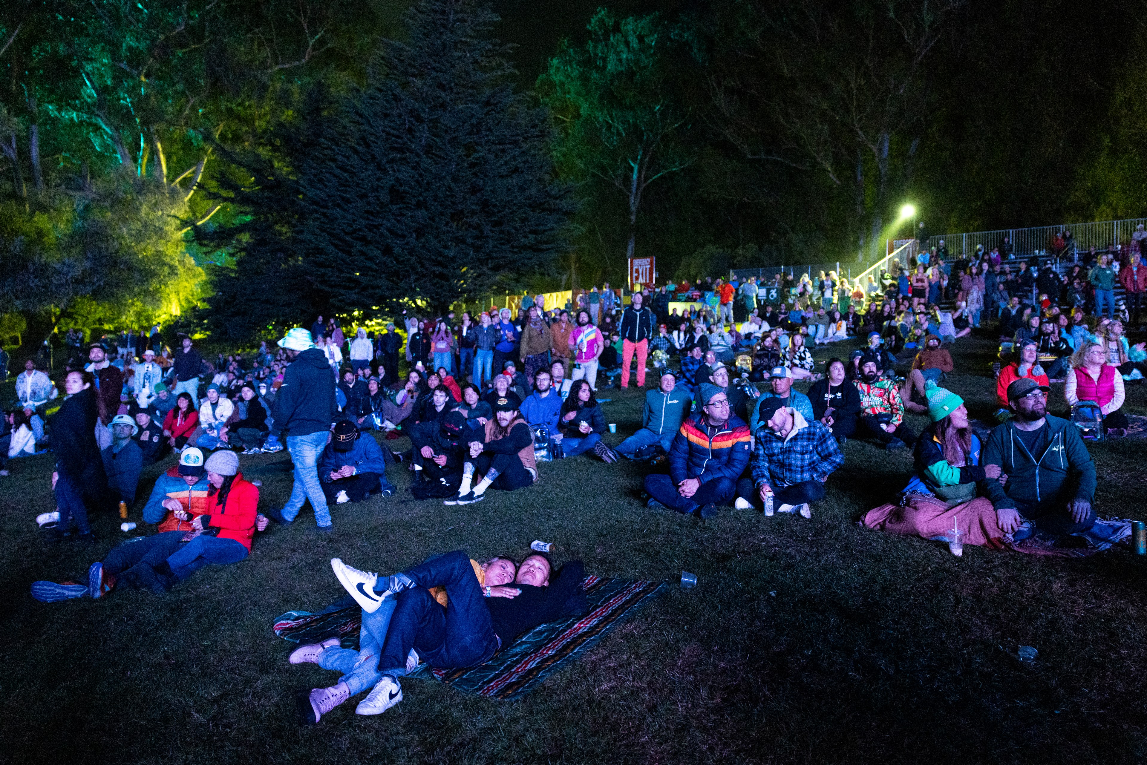 A crowd of people is seated on grass at an outdoor nighttime event, with some lying on blankets and others sitting in groups, surrounded by illuminated trees.