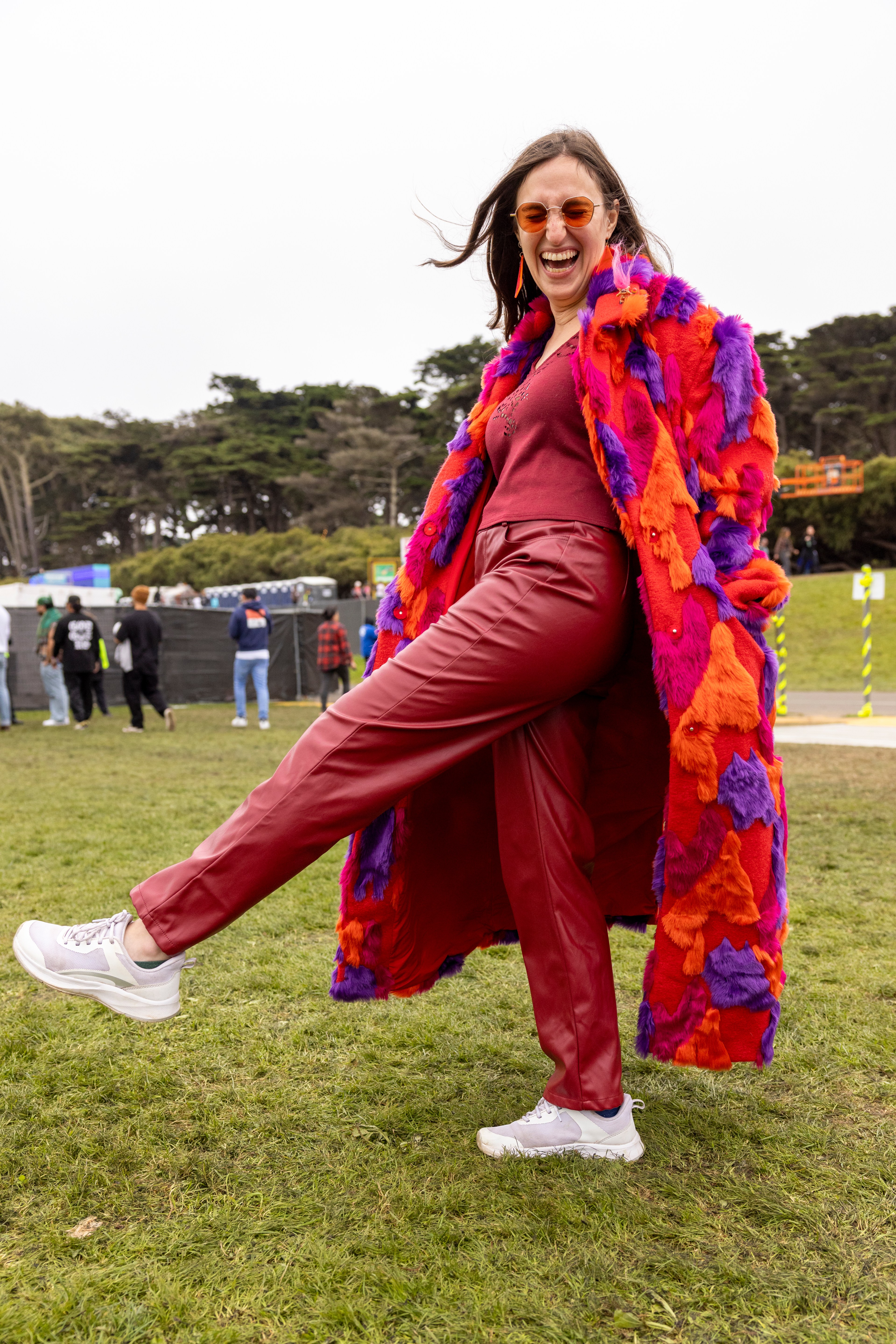 A joyful woman stands on grass, kicking up a leg playfully. She wears orange sunglasses, a vibrant furry coat, red pants, and white sneakers, with trees in the background.