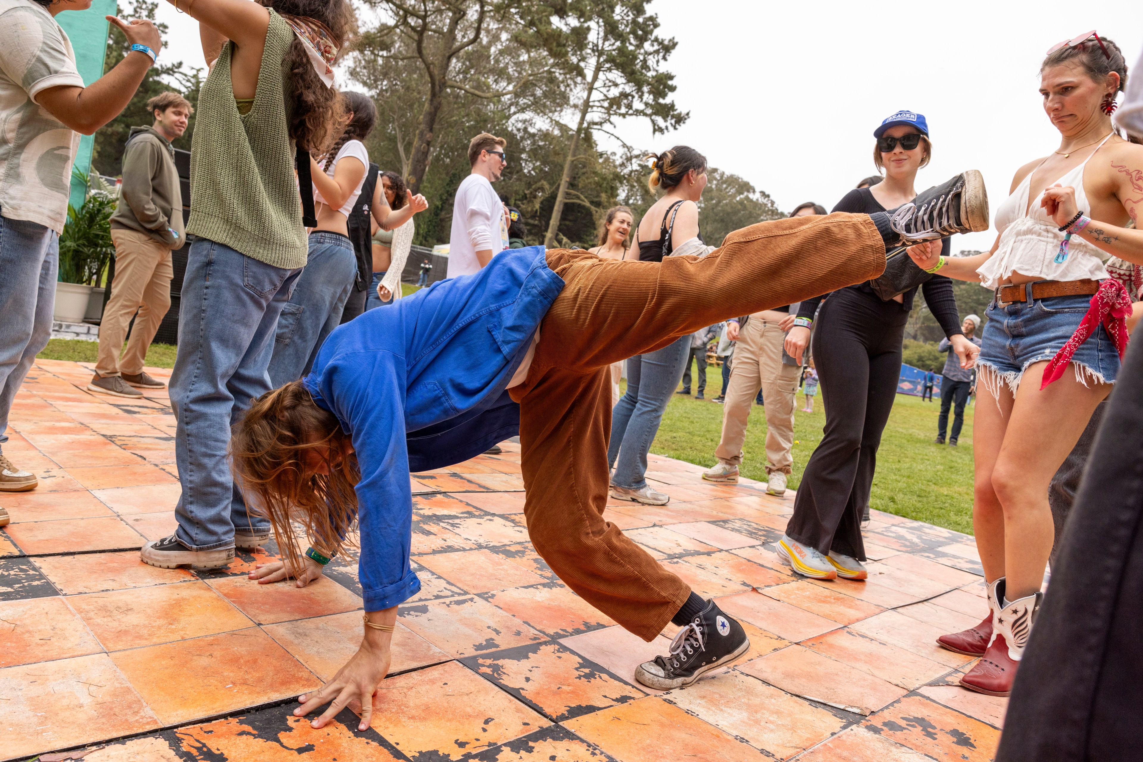 A person in a blue jacket and brown pants does a handstand on an outdoor dance floor while a group of people nearby dance and watch.