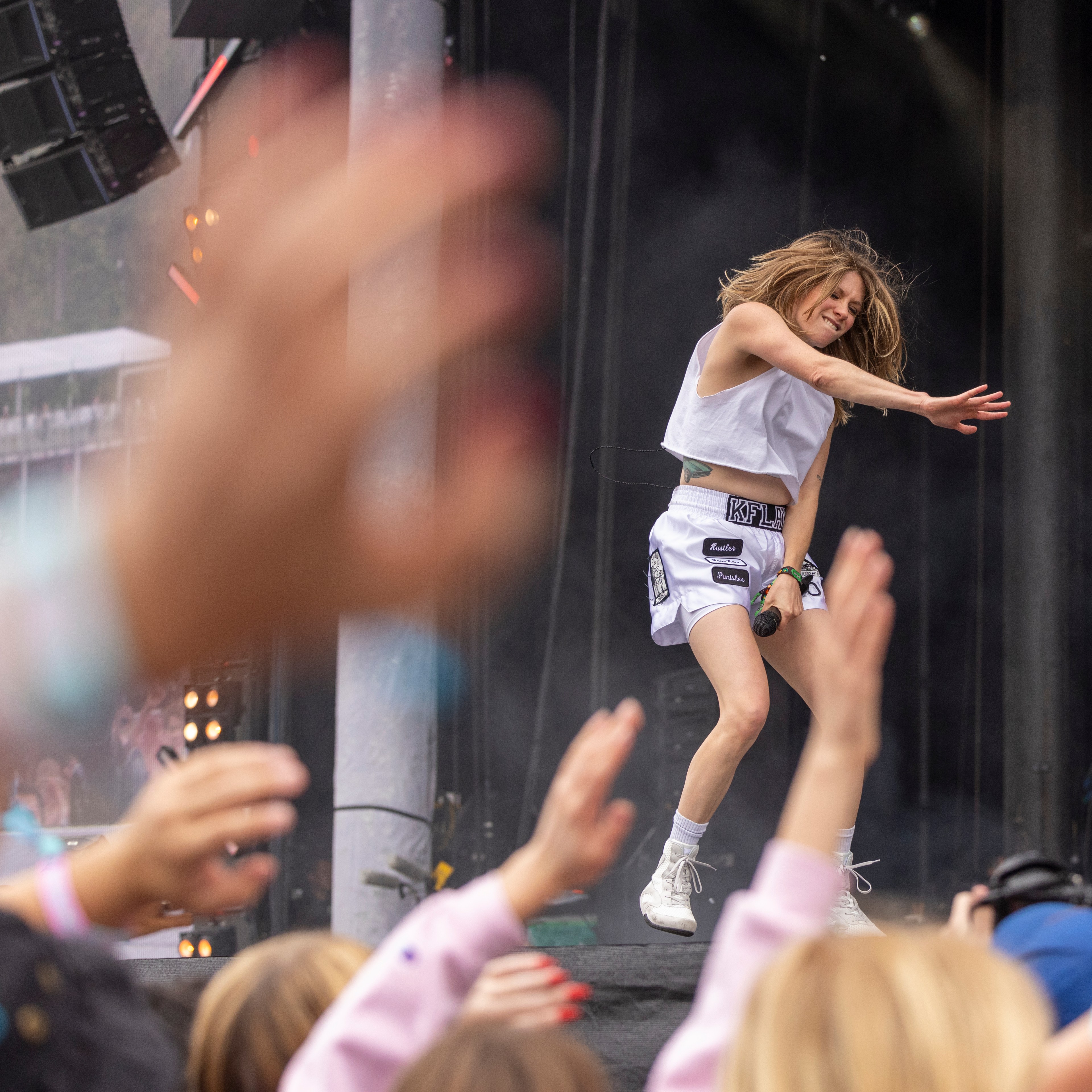 A performer in a white outfit energetically sings on stage. The crowd in the foreground has raised hands, clapping and cheering. Smoke and lighting effects enhance the scene.