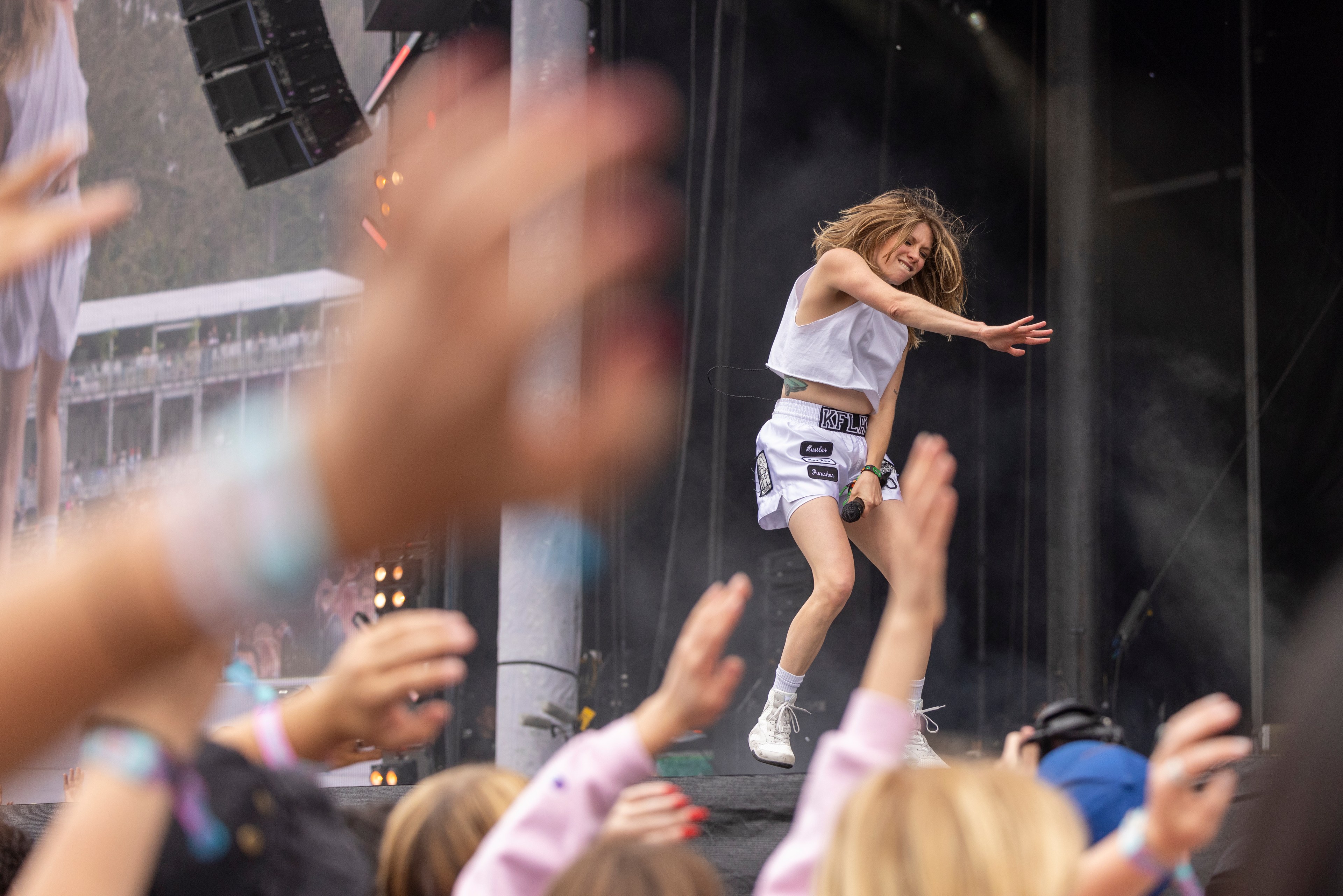 A performer in a white outfit energetically sings on stage. The crowd in the foreground has raised hands, clapping and cheering. Smoke and lighting effects enhance the scene.