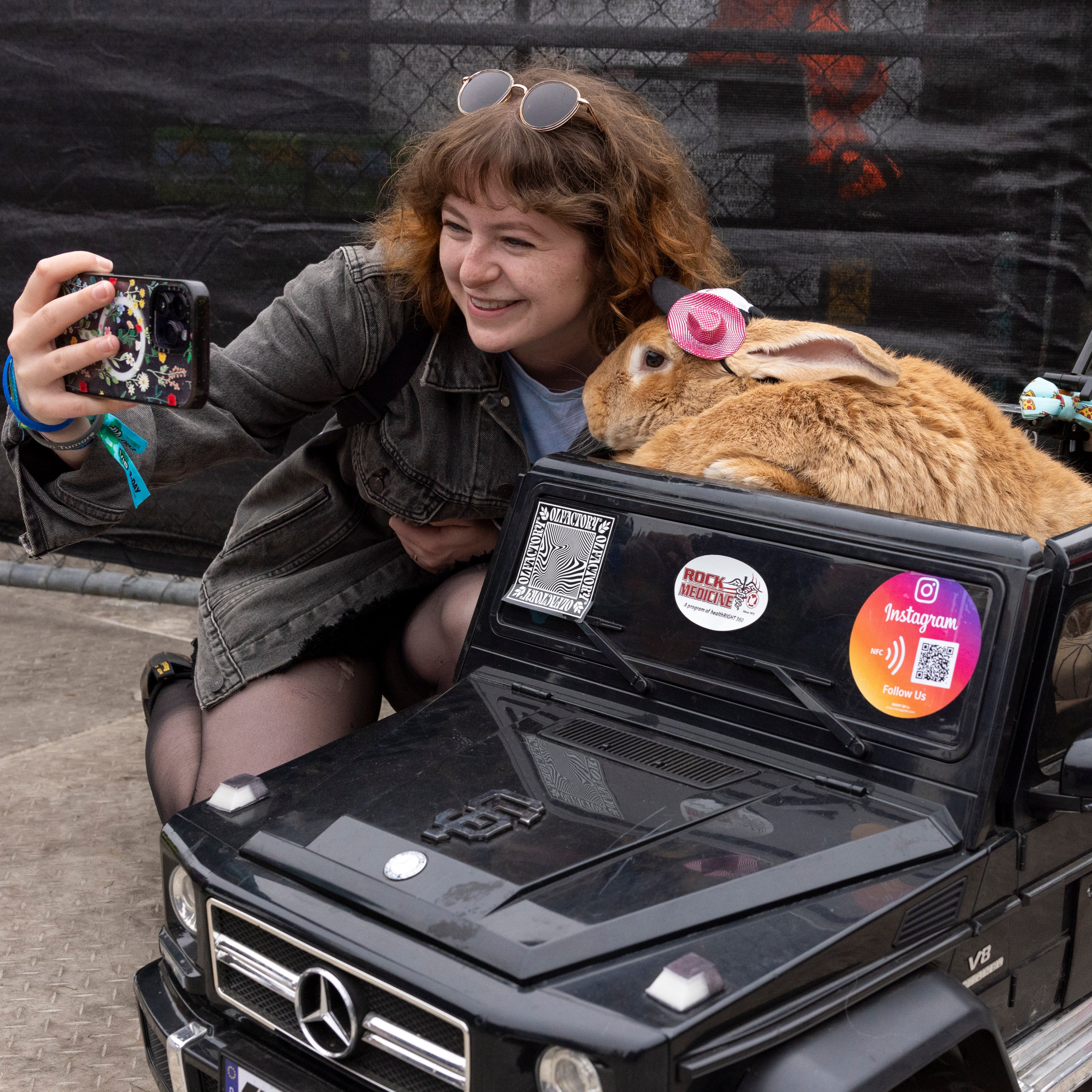 A smiling woman takes a selfie with a large brown rabbit wearing a pink hat, sitting in a black toy Mercedes-Benz car, decorated with various stickers.