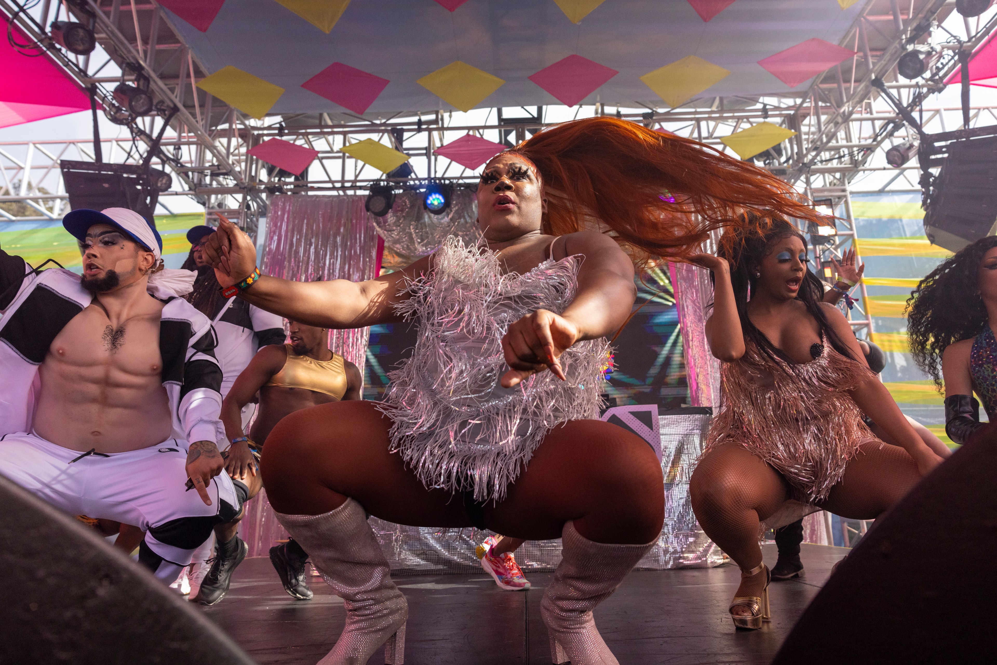 Performers, dressed in elaborate and glittery costumes, dance energetically on stage under a colorful, geometric-decorated canopy with stage lights overhead.