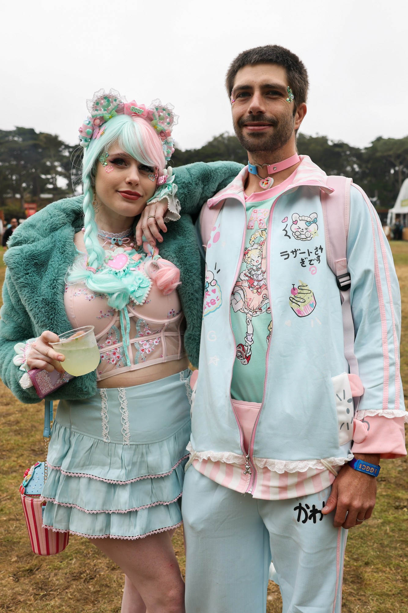 A colorful couple poses at an outdoor event. The woman wears a pastel goth-style outfit with turquoise and pink accents, while the man sports matching pastel attire.