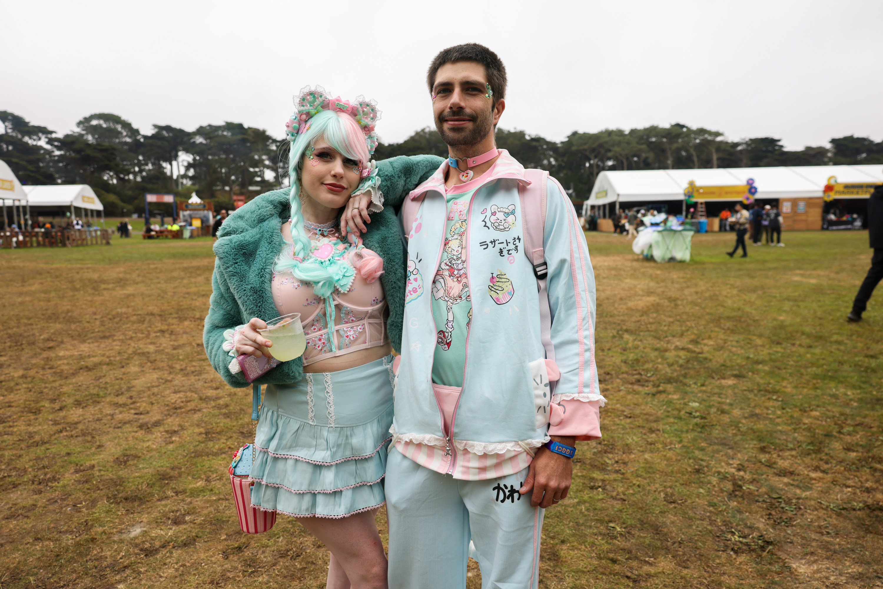 A colorful couple poses at an outdoor event. The woman wears a pastel goth-style outfit with turquoise and pink accents, while the man sports matching pastel attire.