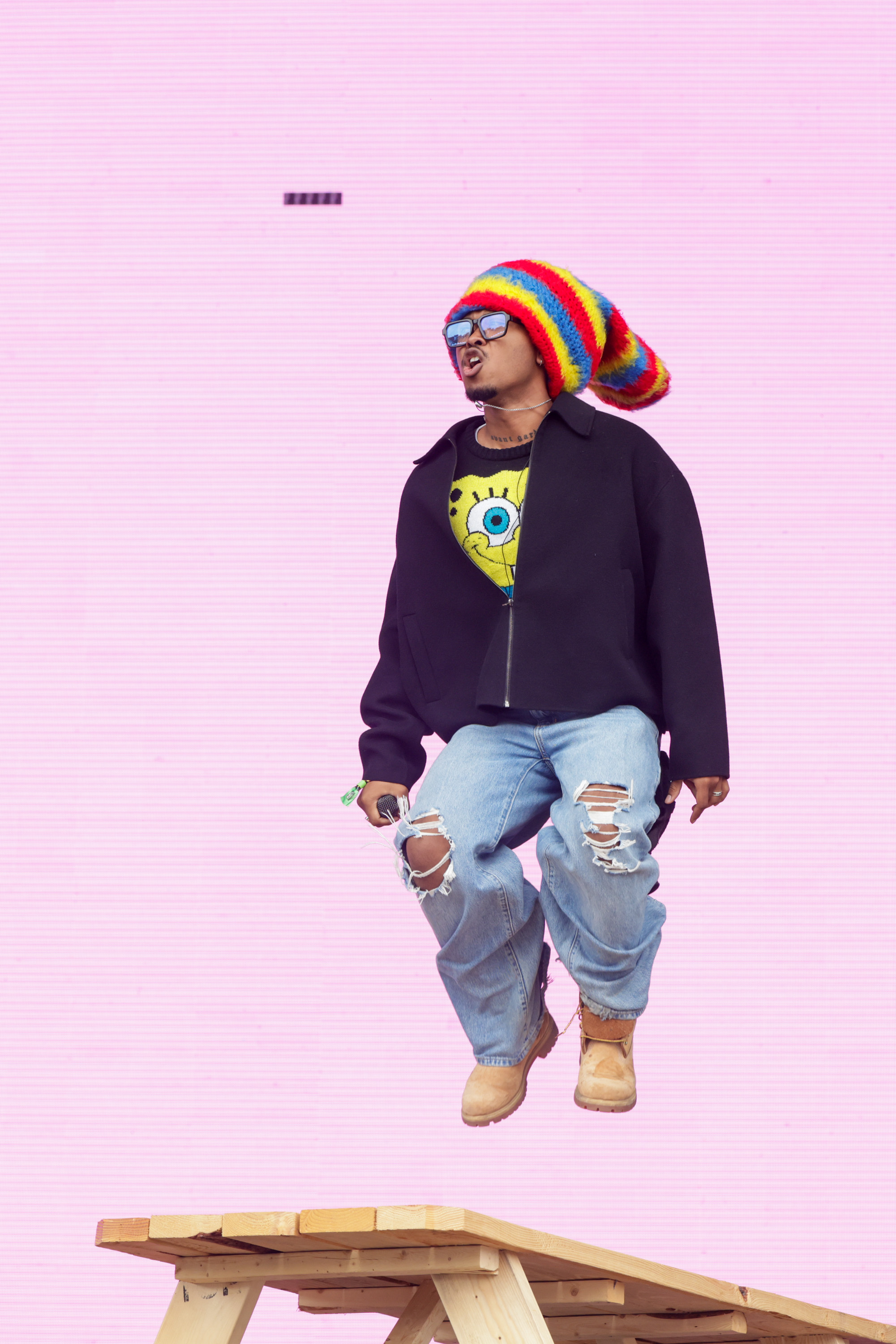 A person with a rainbow beanie, sunglasses, and a Spongebob sweater is jumping on a wooden table against a pink background. They are also wearing a black jacket and ripped jeans.