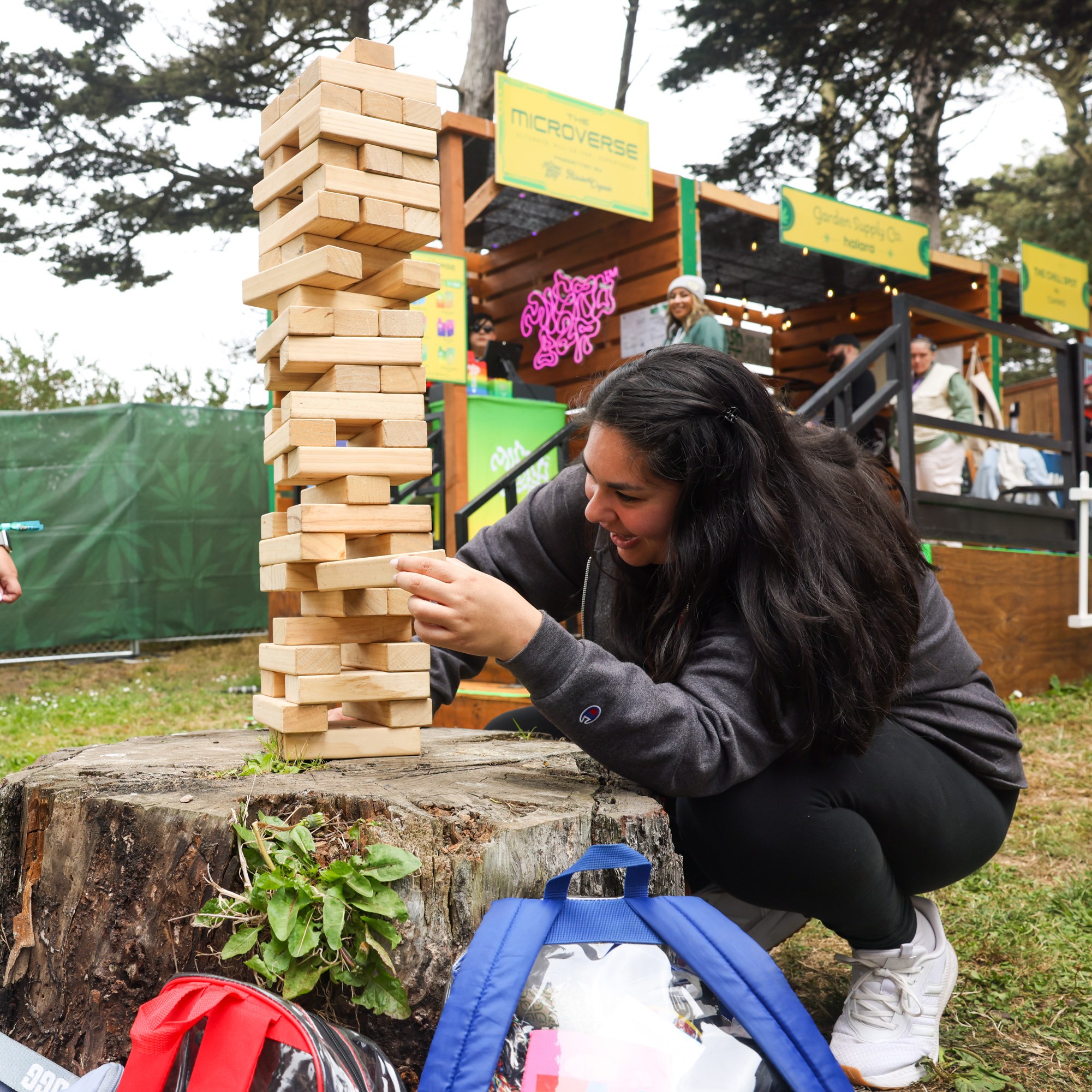 Two people enjoy a large Jenga game on a tree stump. One is pushing a block, while the other laughs. The scene is outdoors, near green fencing and booths.