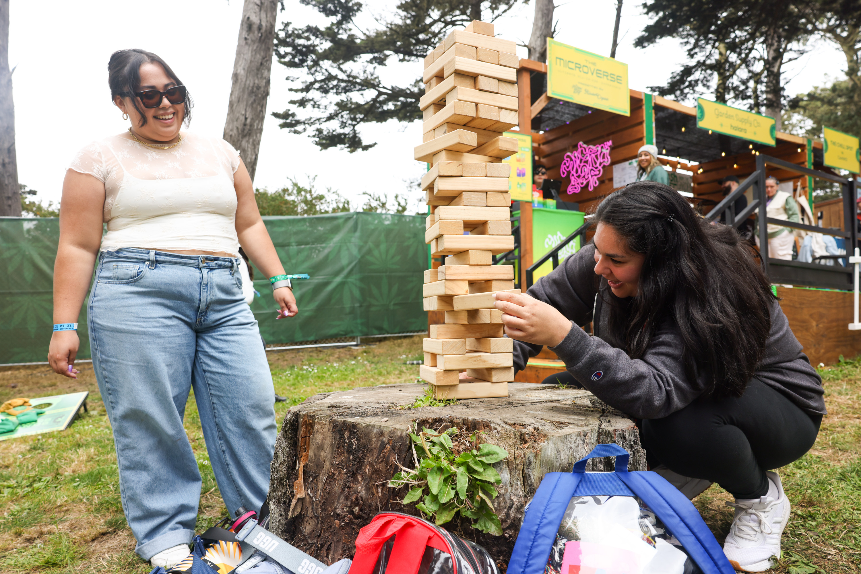 Two people enjoy a large Jenga game on a tree stump. One is pushing a block, while the other laughs. The scene is outdoors, near green fencing and booths.