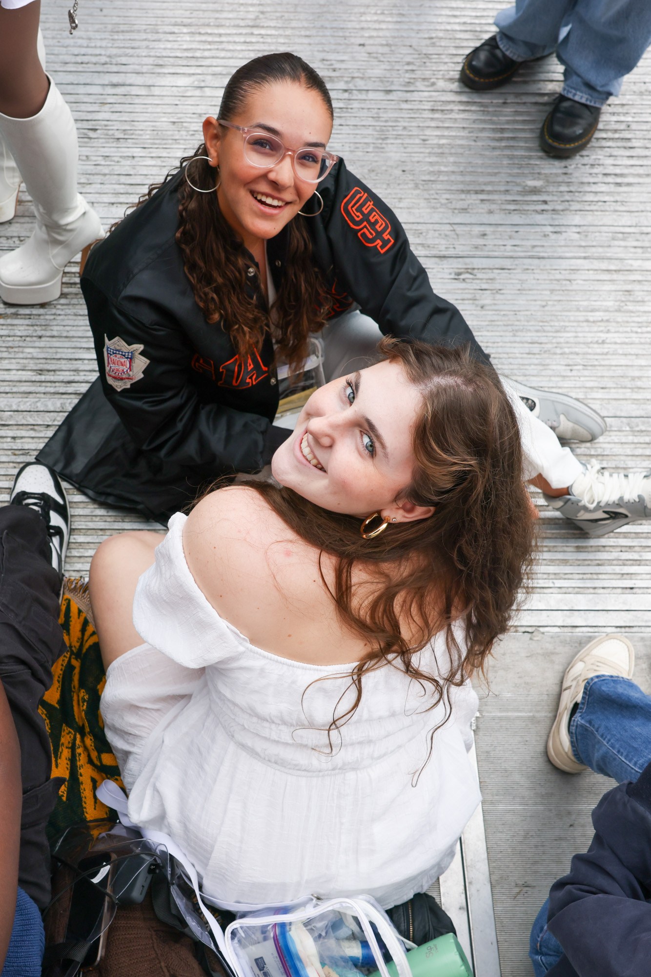 The image shows a group of people sitting on the ground, engaged in various activities. Two women are looking up and smiling towards the camera, while others are using their phones.