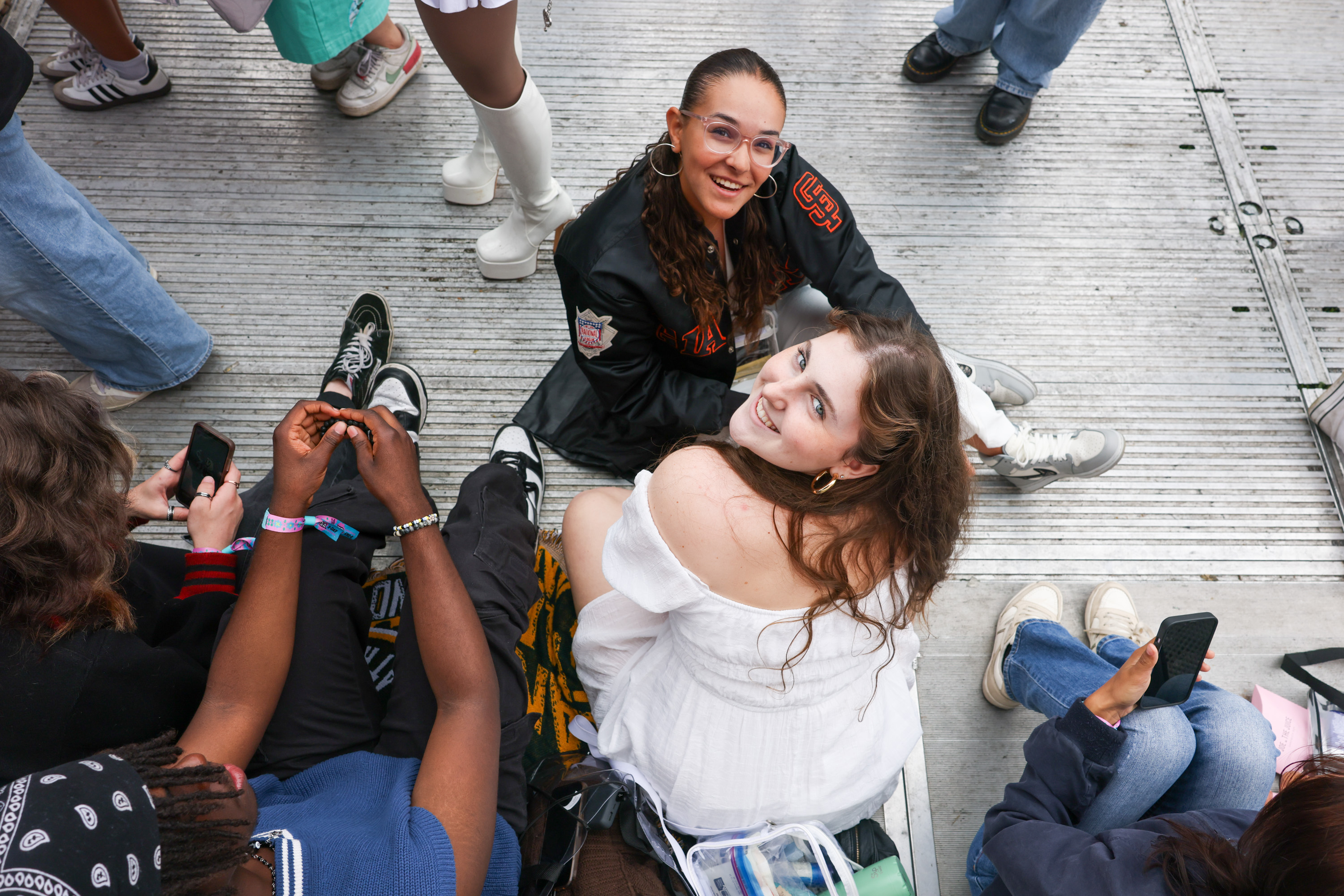 The image shows a group of people sitting on the ground, engaged in various activities. Two women are looking up and smiling towards the camera, while others are using their phones.