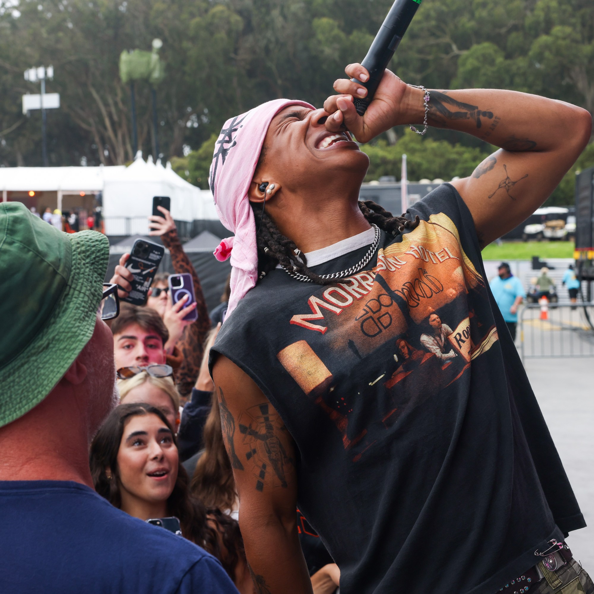 A person in a bandana sings passionately into a microphone while a crowd of fans, some recording with their phones, look on excitedly. One fan is wearing a green hat.