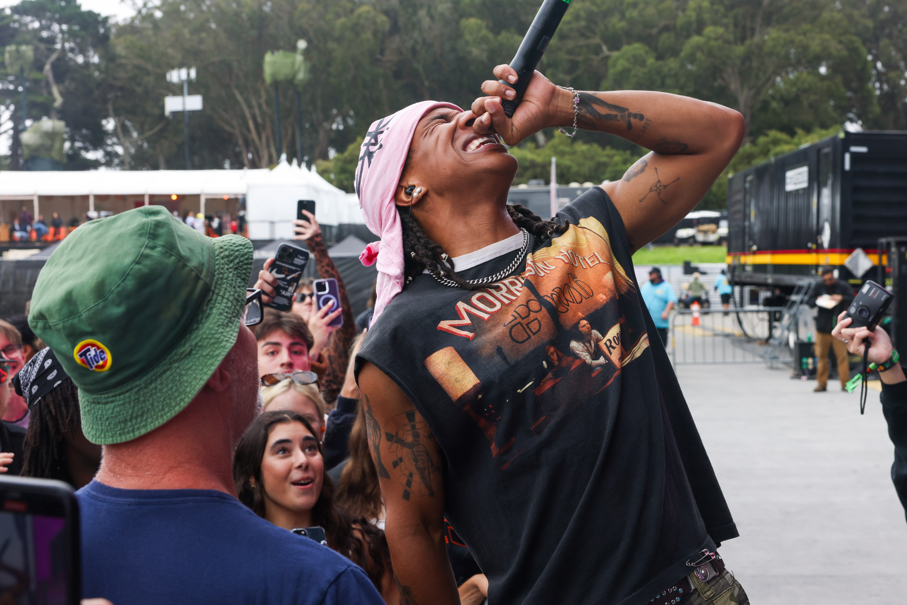 A person in a bandana sings passionately into a microphone while a crowd of fans, some recording with their phones, look on excitedly. One fan is wearing a green hat.