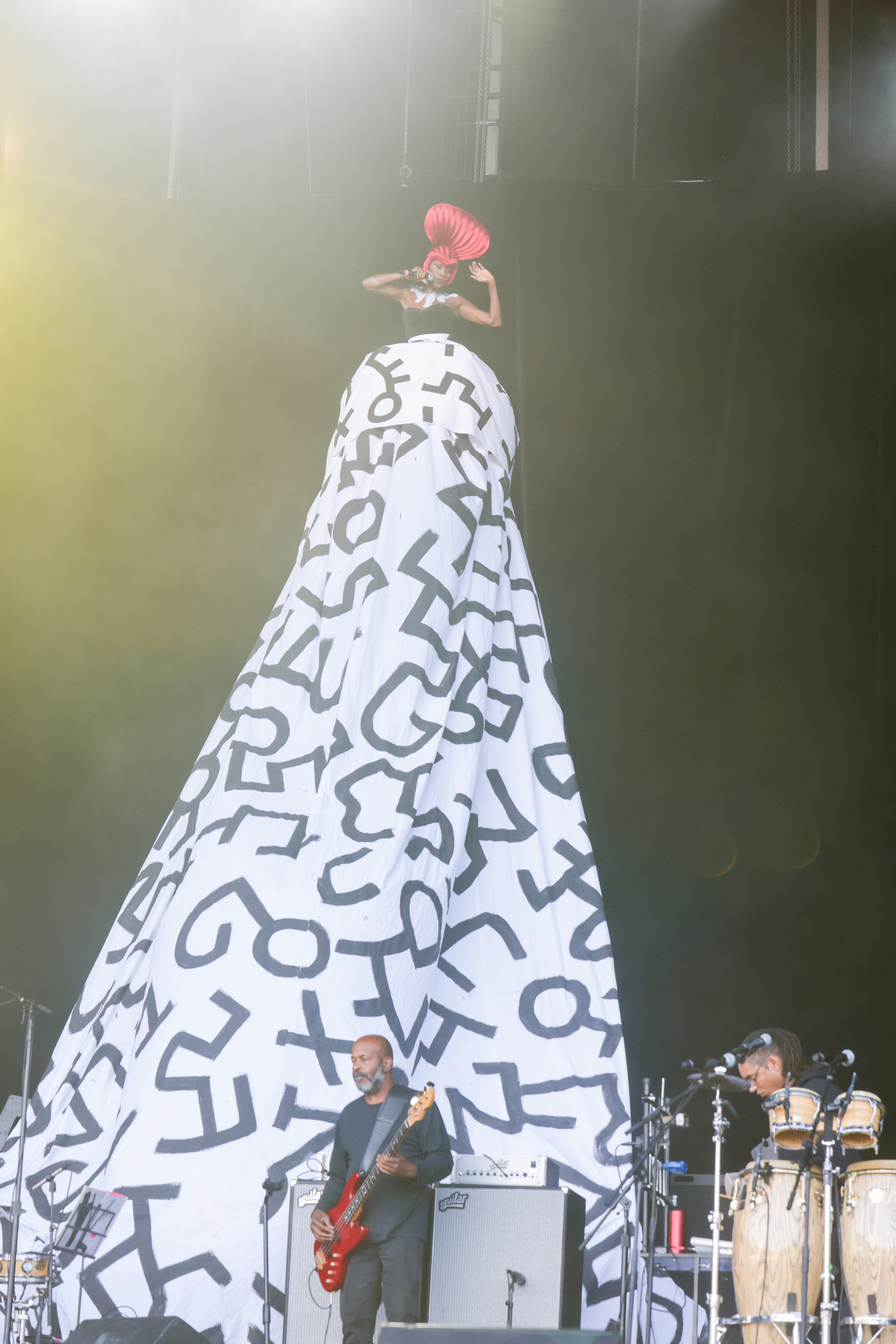 An artist wearing a towering dress with black abstract designs performs on stage with musicians, one playing a guitar and another on drums, set against a dark backdrop.