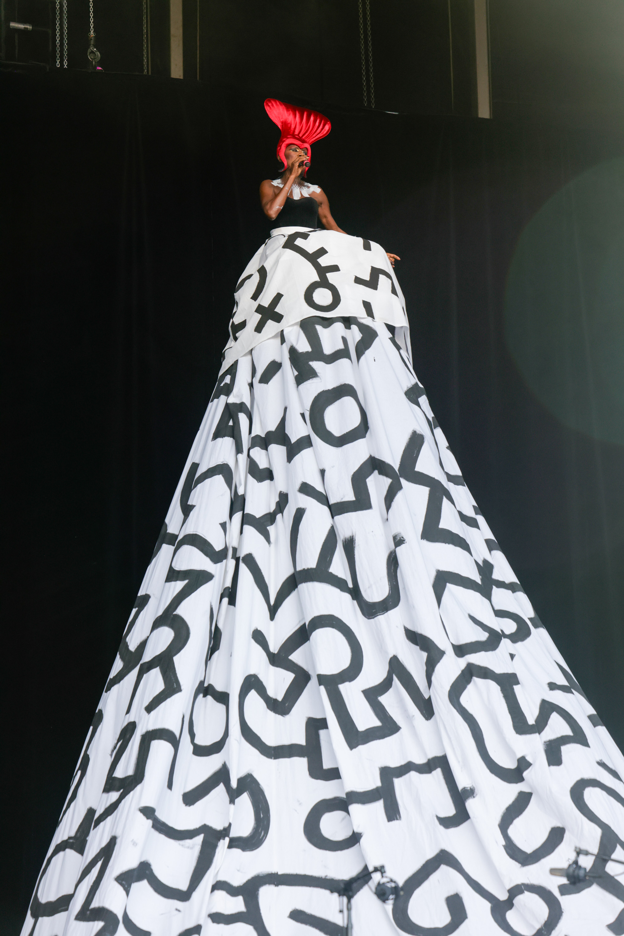 A performer stands tall on a stage, wearing a strikingly large white dress with black abstract symbols and a bright red, tall headpiece, under dark stage lighting.