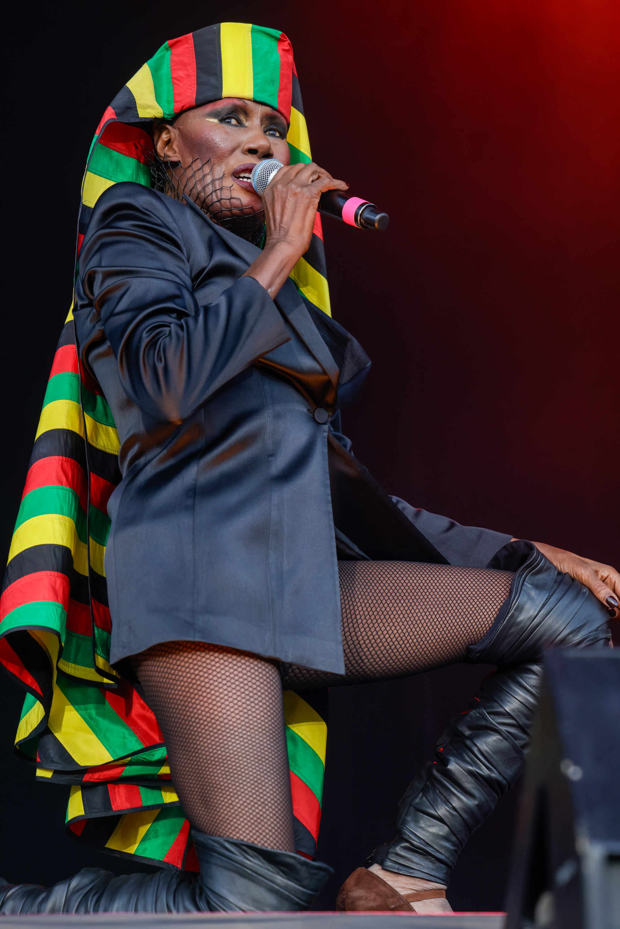 A performer in a sharp black outfit with fishnet stockings and knee-high boots sings into a microphone. They wear a colorful headdress with red, yellow, green, and black stripes.