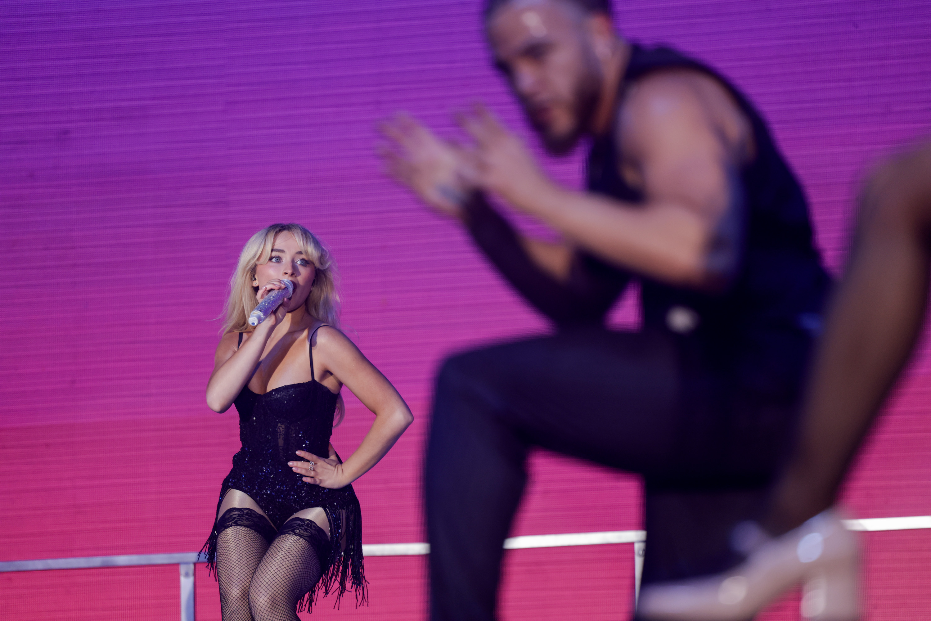 A woman in a black outfit sings into a microphone on stage, with a pink-purple background. A blurred dancer is performing nearby.