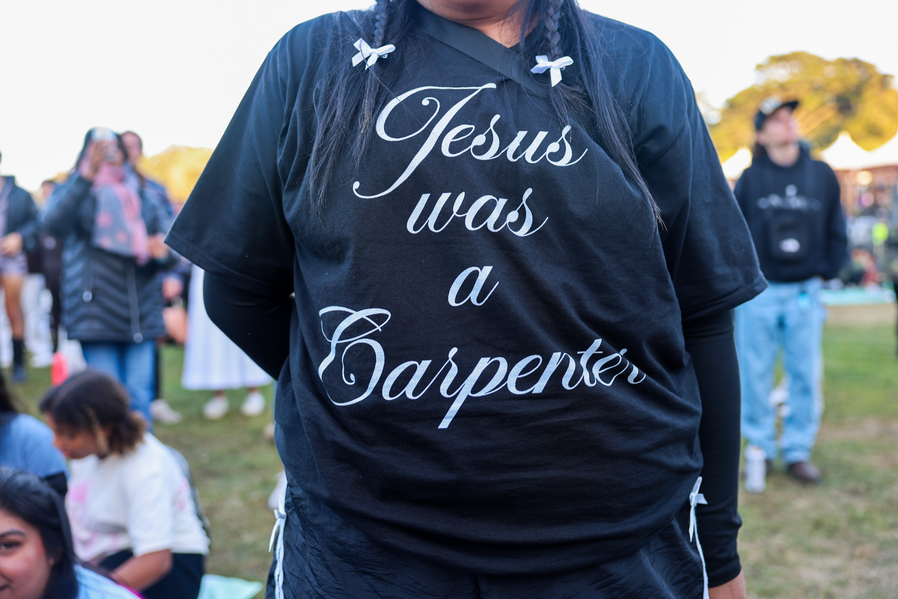 A person wearing a black shirt with the text &quot;Jesus was a Carpenter&quot; stands in an outdoor crowd, with people blurred in the background.