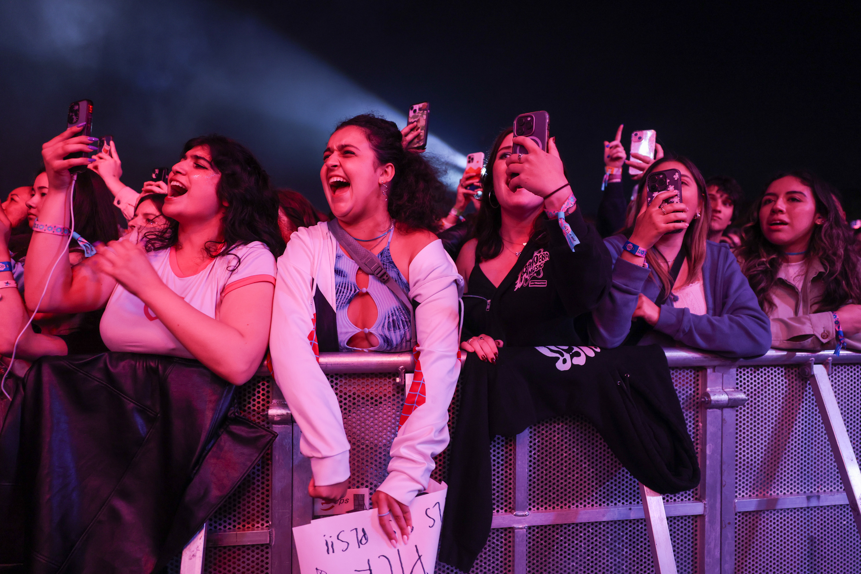 A group of excited concertgoers are pressed against a barrier, some with phones raised to capture the moment, all seemingly enjoying the live performance.